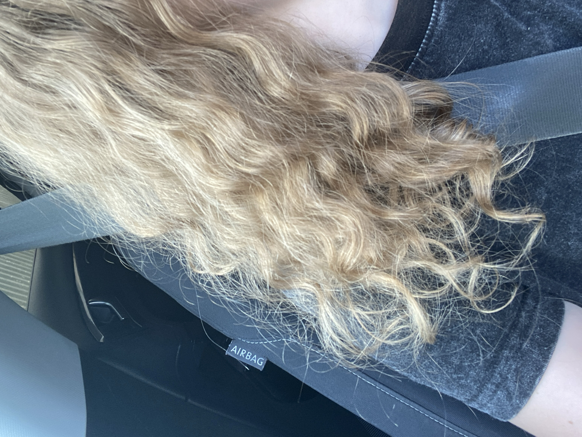 A close-up, eye-level shot shows the side of a person's head and shoulder, with their wavy blonde hair falling forward. The person is wearing a dark gray t-shirt with a textured pattern. They are sitting in a car, indicated by a gray seatbelt crossing their chest and a visible black car seat with an "AIRBAG" label. The background is blurred, but suggests the interior of a vehicle. The lighting is natural and bright.