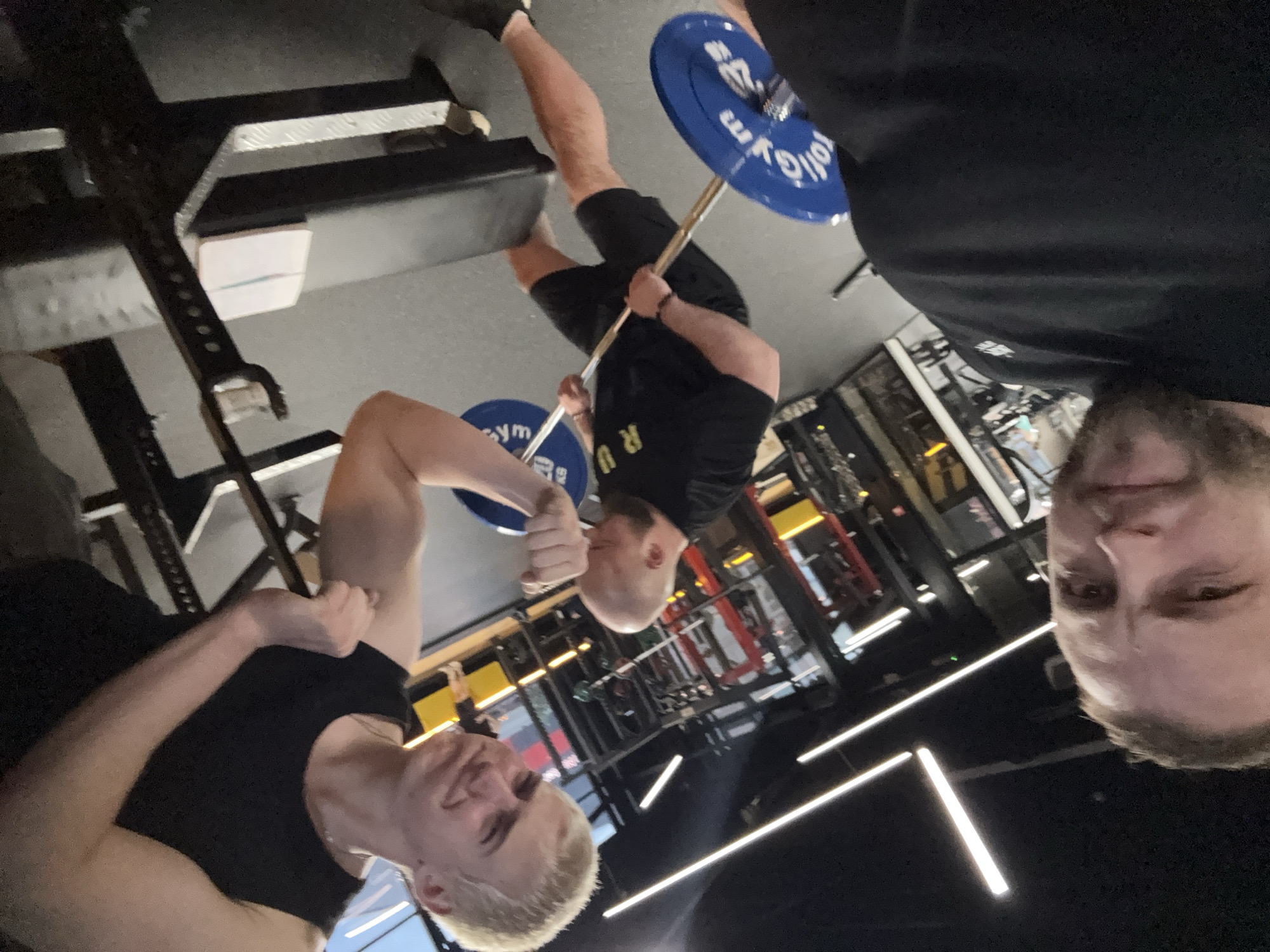 The image, rotated 90 degrees counter-clockwise from its upright orientation, captures three men in what appears to be a gym. The perspective suggests a selfie.
In the foreground, on the right side of the rotated image, a man with a beard and dark clothing is partially visible, looking towards the camera. To the left in the foreground, another man with light-colored hair, wearing a black tank top, is flexing his right bicep and smiling slightly.
In the midground, a third man with a bald or shaved head, wearing a black t-shirt with yellow letters "R U" and black shorts, is holding a barbell. The barbell is fitted with blue weight plates that display the word "gym" and what appears to be part of a brand name ("ollo") in reverse. He seems to be in the middle of an exercise, with his left arm flexed.
The background reveals typical gym equipment, including weight racks, benches, and other machinery. The setting features dark floors and walls, illuminated by bright, linear LED light fixtures on the ceiling. The overall scene depicts friends working out together in a modern fitness center.