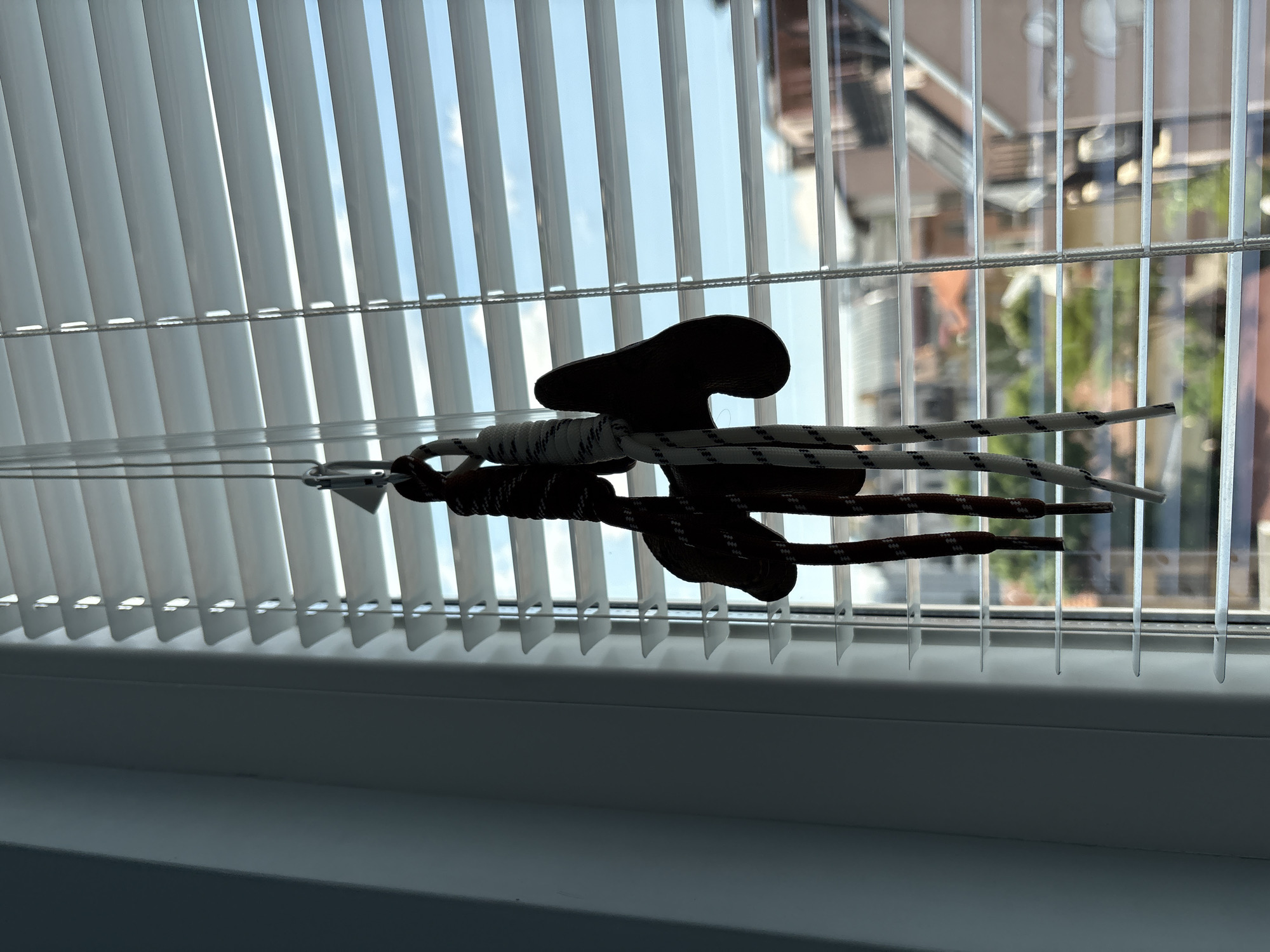 The image presents a detailed view of two shoelaces suspended in front of a window, creating a strong silhouette against the bright outdoor light. The shoelaces consist of one dark, solid-colored strand and one white strand with black flecks, both hanging vertically. They are tied to two distinct, dark, silhouetted objects, which are themselves attached to a silver-colored carabiner or clip at the top.
The interior environment on the left features a light-colored wall or window frame, cast in shadow, which transitions into a bright white horizontal Venetian blind system covering the window. The blinds are partially open, allowing several horizontal strips of natural light to penetrate the room.
Through the gaps in the blinds, an outdoor urban scene is visible under a clear, bright blue sky with sparse white clouds. The view encompasses various buildings, indicating a city setting. Notable architectural elements include a prominent reddish-brown tiled roof on one building, suggestive of traditional European construction, and other structures with more muted, possibly lighter-colored facades. Hints of green foliage are visible, suggesting trees or other landscaping within the urban fabric. This composite view of buildings and sky is consistent with an urban residential or commercial area in Sofia, Bulgaria, observed from an elevated perspective.