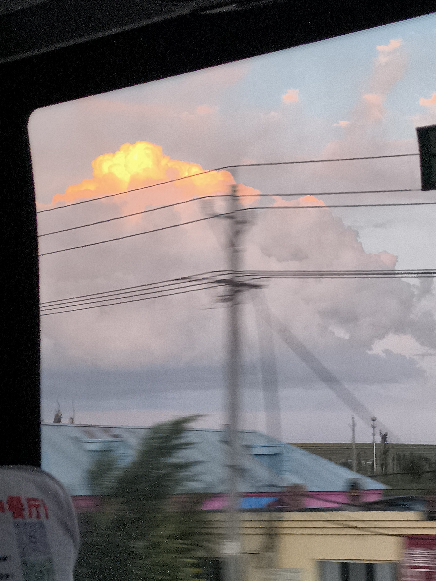 The image depicts a view from a window, likely a bus or car, looking out at a landscape during what appears to be sunset. The sky is filled with soft pink and blue hues, with a prominent bright orange cloud formation dominating the upper left quadrant. Several horizontal power lines crisscross the scene, creating a sense of depth and obscuring parts of the sky and landscape. In the mid-ground, a telephone pole with wires extends vertically. Below the sky, there are rooftops of buildings, some with light blue and pink colored surfaces, and blurred green foliage. The overall impression is one of a transient view, captured from a moving vehicle, with the vibrant colors of the sky suggesting a picturesque moment. The location is identified as Guozigou Pasture, China, though this is not visually discernible from the image itself.