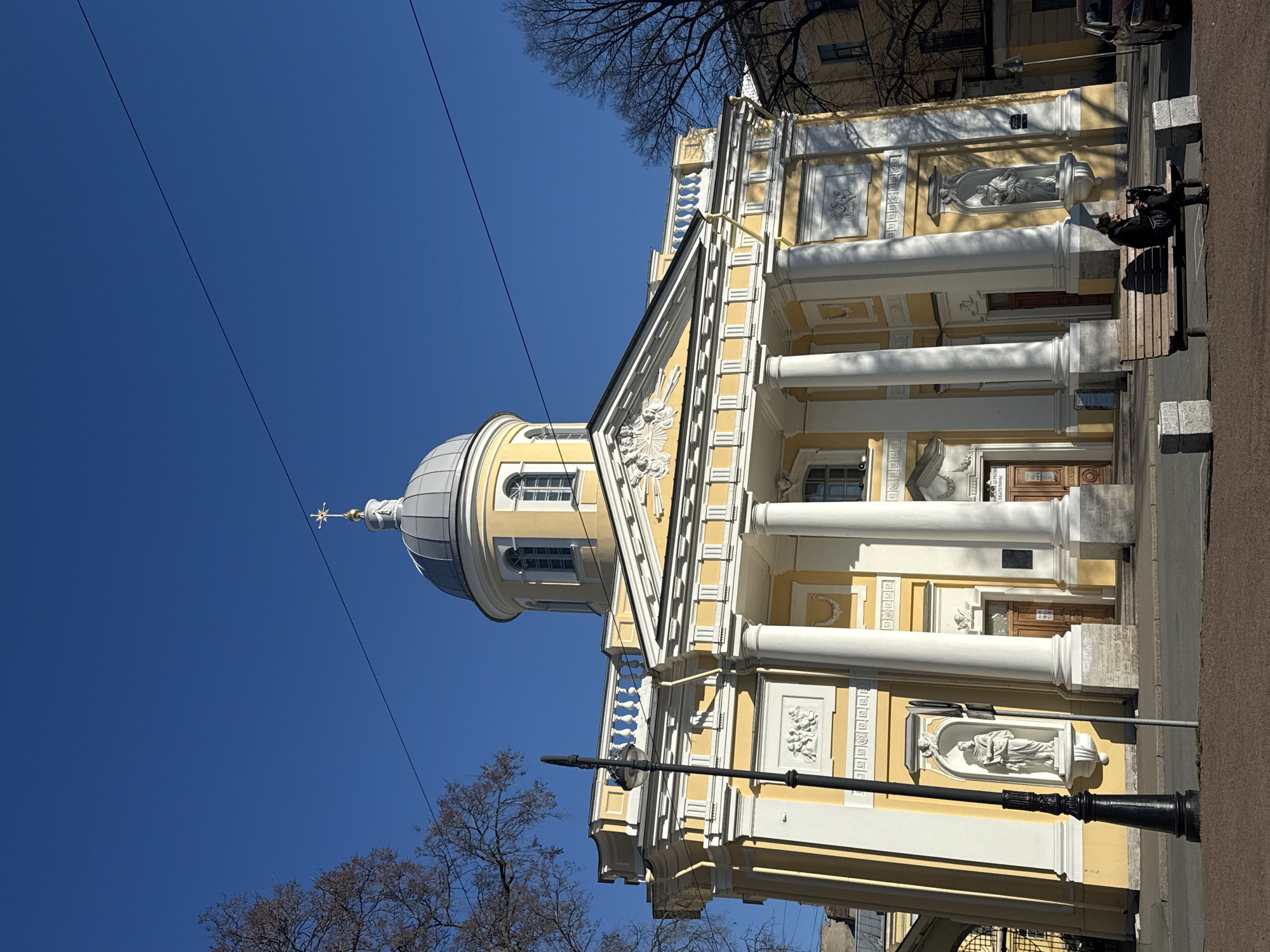 The image depicts the Church of St. Catherine on Vasilyevsky Island in Saint Petersburg, Russia. The yellow and white neoclassical building features a dome, columns, and sculptural details. A person sits on a bench near the building. Power lines are visible against a clear blue sky. Bare trees are present in the foreground and background. A streetlamp is also visible in the lower part of the image.