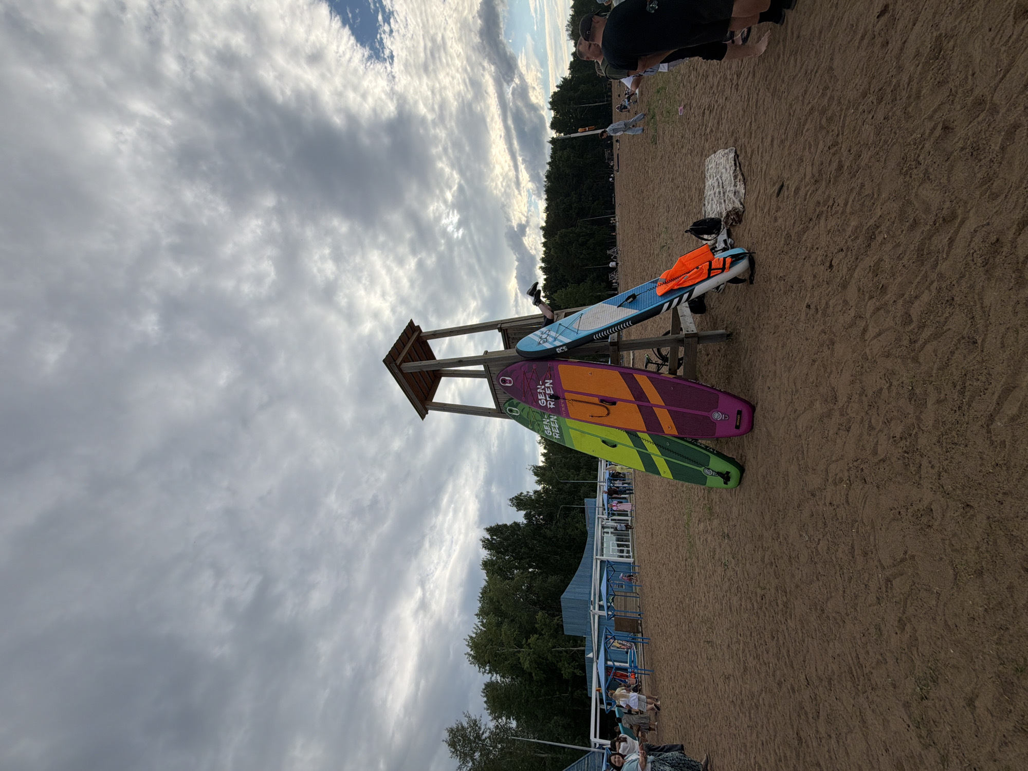 This is a photograph taken on a beach, likely in Chaikovsky, Russia, given the context. The image shows several people on a sandy beach, with a backdrop of trees and a cloudy sky. The time of day appears to be late afternoon or early evening, judging by the light and the cloudy weather.
In the foreground, there are three stand-up paddleboards stacked on a wooden structure. One paddleboard is blue with white accents and an orange life vest on it. Another is purple with yellow and green accents, and the third is green with yellow accents. Visible text on the purple paddleboard reads "GEN GEN-RLEN-RLEN".
Further back on the beach, a group of people are visible, some sitting and others standing. A few are near a set of blue structures that appear to be part of a recreational area or beach facilities. The overall atmosphere seems relaxed, with people enjoying the beach. The clouds are dense and cover most of the sky, suggesting overcast conditions.