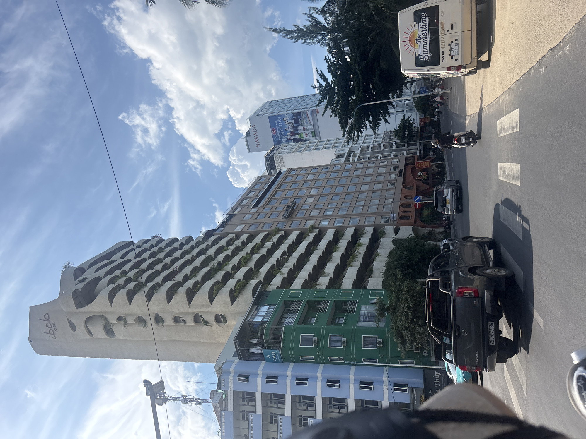 A street scene in Nha Trang, Vietnam, depicts a multilane road under a partly cloudy sky. On the left, a tall, distinctive cream-colored hotel with balconies shaped like upturned petals, labeled "ibalo HOTEL," dominates the skyline. Next to it, a smaller building is painted in vibrant blue with numerous windows. Adjacent to the blue building is a five-story green structure with white-framed windows and balconies. Further down the street, a modern white skyscraper with a large advertisement is visible. The road itself features a crosswalk and various vehicles, including a dark gray Nissan Navara pickup truck with the license plate "29C-877.84" stopped at the crosswalk. A person on a scooter is also visible on the road, as is a white van with "The Summertime" printed on its side and license plate "49D-004.18." Trees line the right side of the road, and a streetlamp is present. The overall impression is a bustling urban environment on a sunny day.