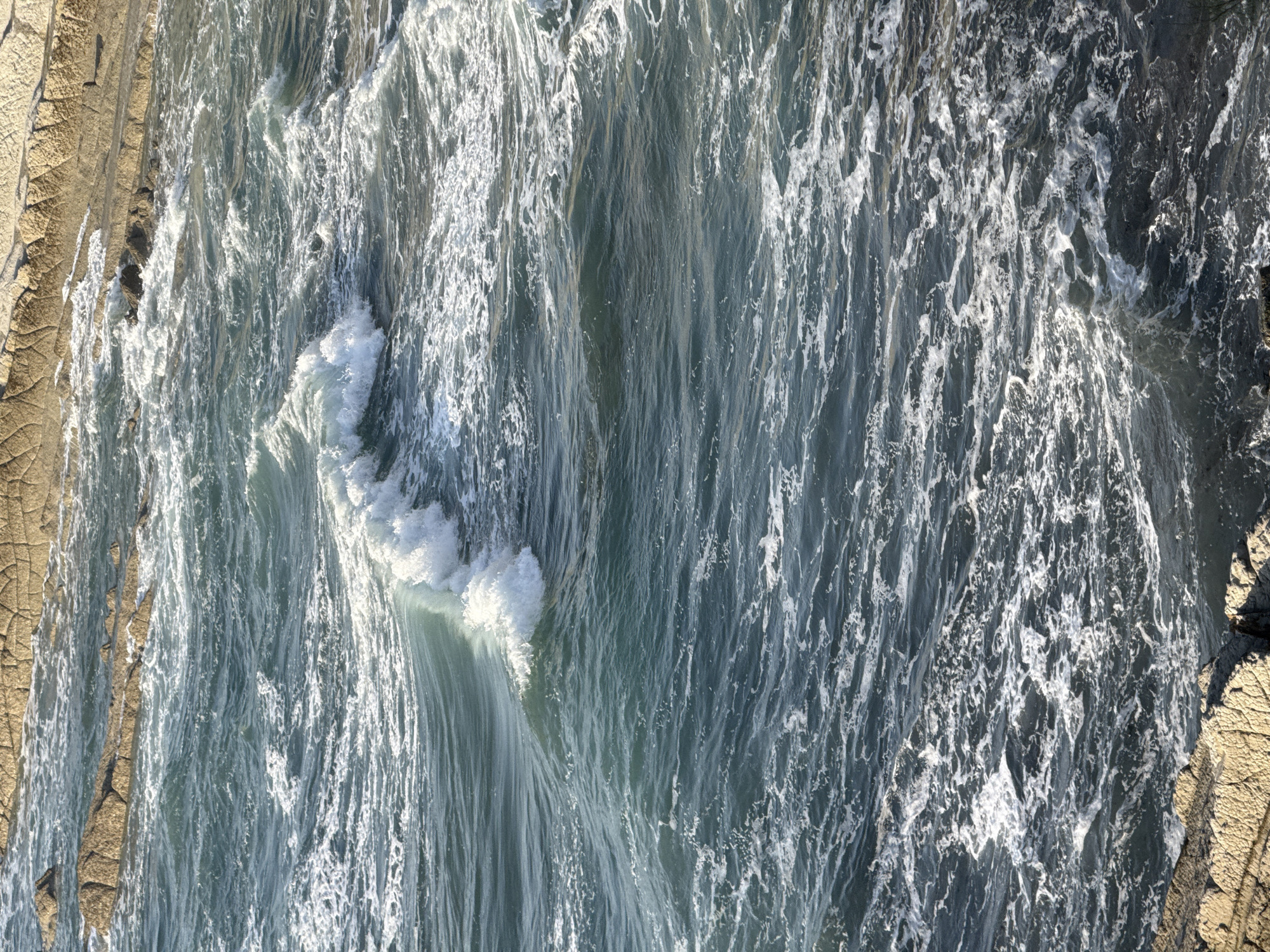 This is a high-angle, close-up shot of the turbulent ocean waters near the coast of Sopela, Spain. The water is a deep teal color, churned into white foam by the crashing waves. The waves are dynamic, with a prominent crest of white water breaking in the mid-left of the frame, and smaller whitecaps scattered throughout the rest of the image. The sun glints off the water's surface, creating highlights. In the upper portion of the image, weathered, textured rock formations, likely part of the Sopela coastline, are visible. The rocks are a light tan color and show signs of erosion. The overall impression is one of natural, untamed coastal energy.