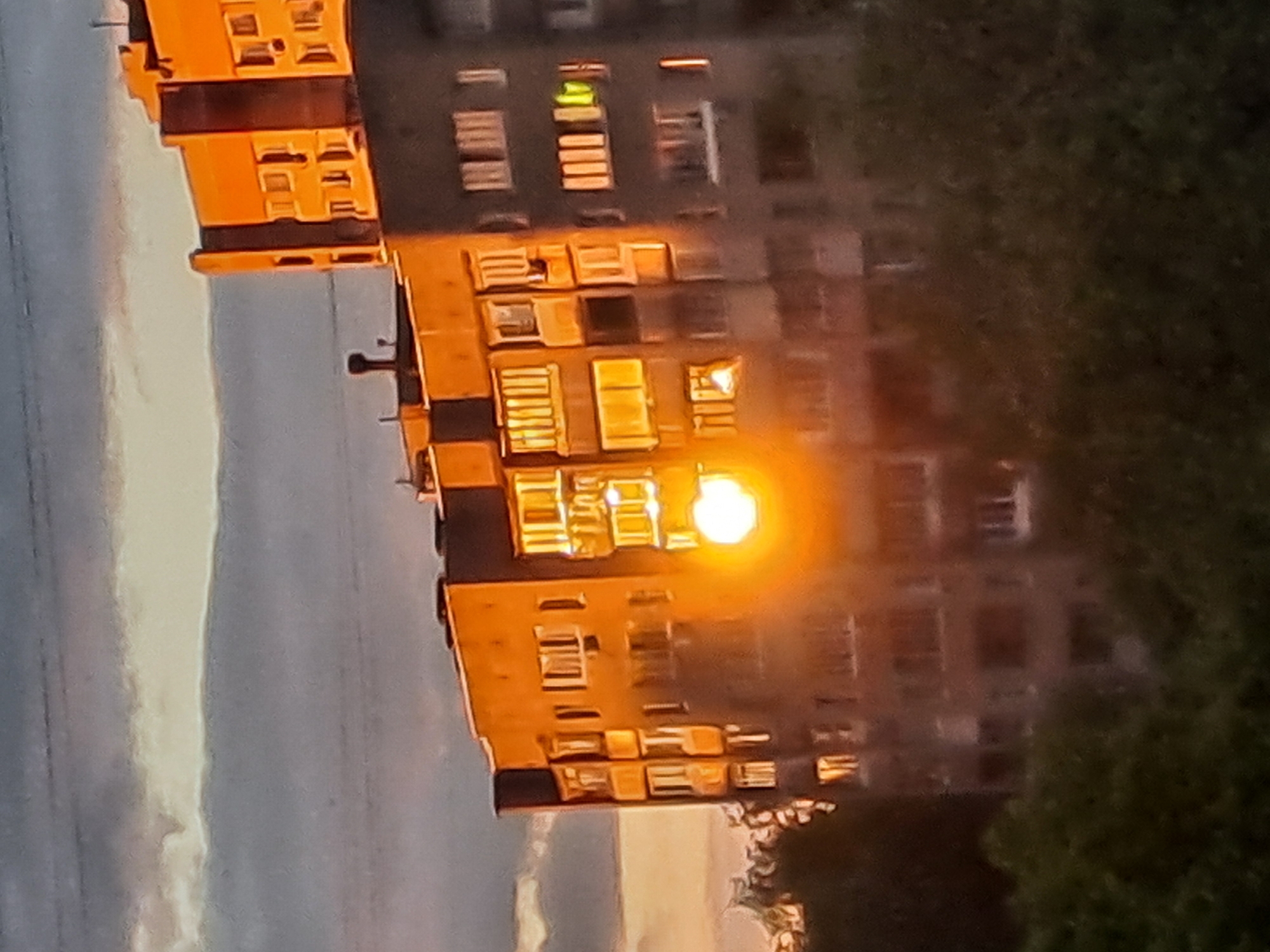 The image shows a tall residential building in Elektrostal, Russia, with many windows. Some windows are illuminated with a bright orange light, possibly from interior lighting. The building appears to be multi-storied. To the right of the building is a green, leafy tree. To the left is a sky with pale colors.