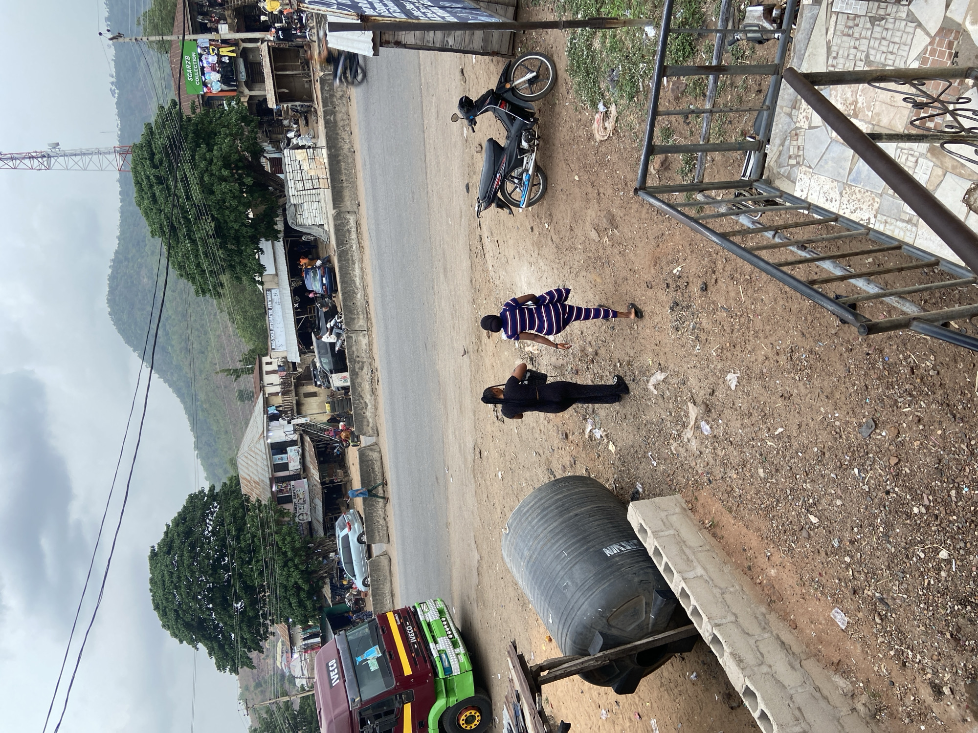 The image captures a roadside scene in Lokoja, Nigeria, under an overcast sky, with lush green hills forming the backdrop. The view is from an elevated position, looking down onto a paved road and its adjacent dirt shoulders.
In the foreground, a large black water tank, partially visible concrete blocks, and metal stairs are situated on the dusty ground, which is littered with some debris. Two individuals are present on the dirt shoulder near the road. One person, dressed in a blue and white striped outfit, walks away from the camera. Nearby, another person in a dark, possibly black, outfit stands facing the road, seemingly engaged with something in their hands. A black motorcycle is parked on the dirt shoulder.
The paved road features light traffic, with several cars parked along its side. A distinctive red and green IVECO truck is partially visible in the left foreground. Buildings line the road, suggesting a commercial area. Visible signs include "SCARZIB COLLECTION" on a green banner above one shop and "MAC YOUNG INTEGRATED MOTORS" on a white sign. Overhead power lines crisscross the sky. A prominent large tree with dense green foliage stands near the road, and further in the background, a cellular tower rises against the sky, preceding the distant green hills.
The scene depicts everyday life in a Nigerian town, with a mix of commercial activity, vehicles, and pedestrians, set against a natural, hilly landscape. The time of day appears to be daylight, possibly early morning or late afternoon given the soft lighting and overcast conditions.