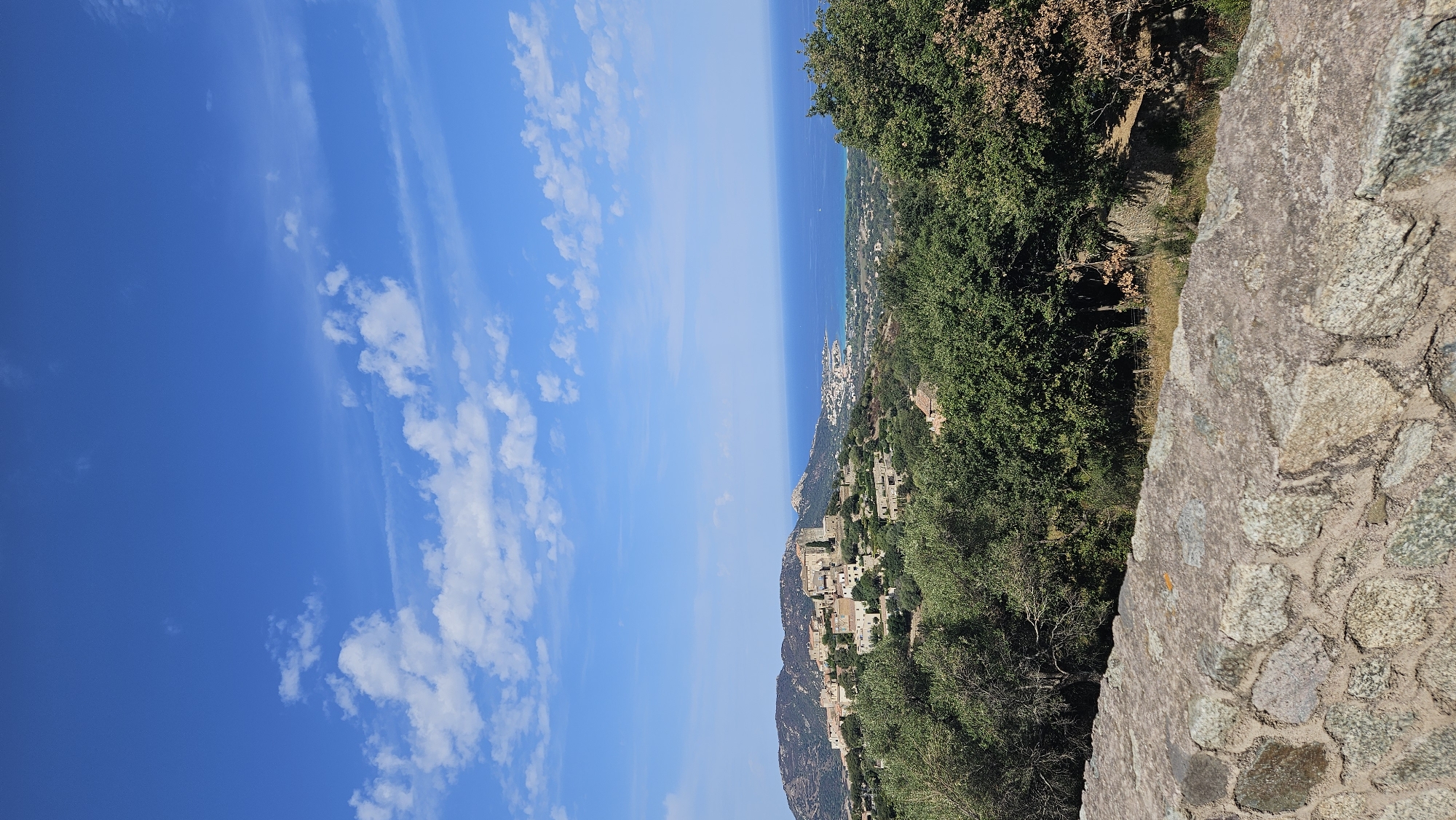 The image depicts a scenic view from a high vantage point overlooking the town of Pigna, France, and the Mediterranean Sea. In the foreground, a textured stone wall made of irregular grey and pinkish stones forms a boundary. Beyond the wall, lush green trees and foliage dominate the middle ground, obscuring parts of the landscape.

In the background, the town of Pigna is visible, nestled on a hillside. The buildings are characterized by their terracotta roofs and light-colored walls, some of which appear to be stone. The town is spread across the slope, with some structures appearing older and more rustic than others. To the right of the town, the blue expanse of the Mediterranean Sea stretches to the horizon, meeting a clear blue sky dotted with scattered white clouds. The sky is a prominent feature, occupying the upper portion of the image. The overall impression is one of a serene coastal village bathed in bright daylight.