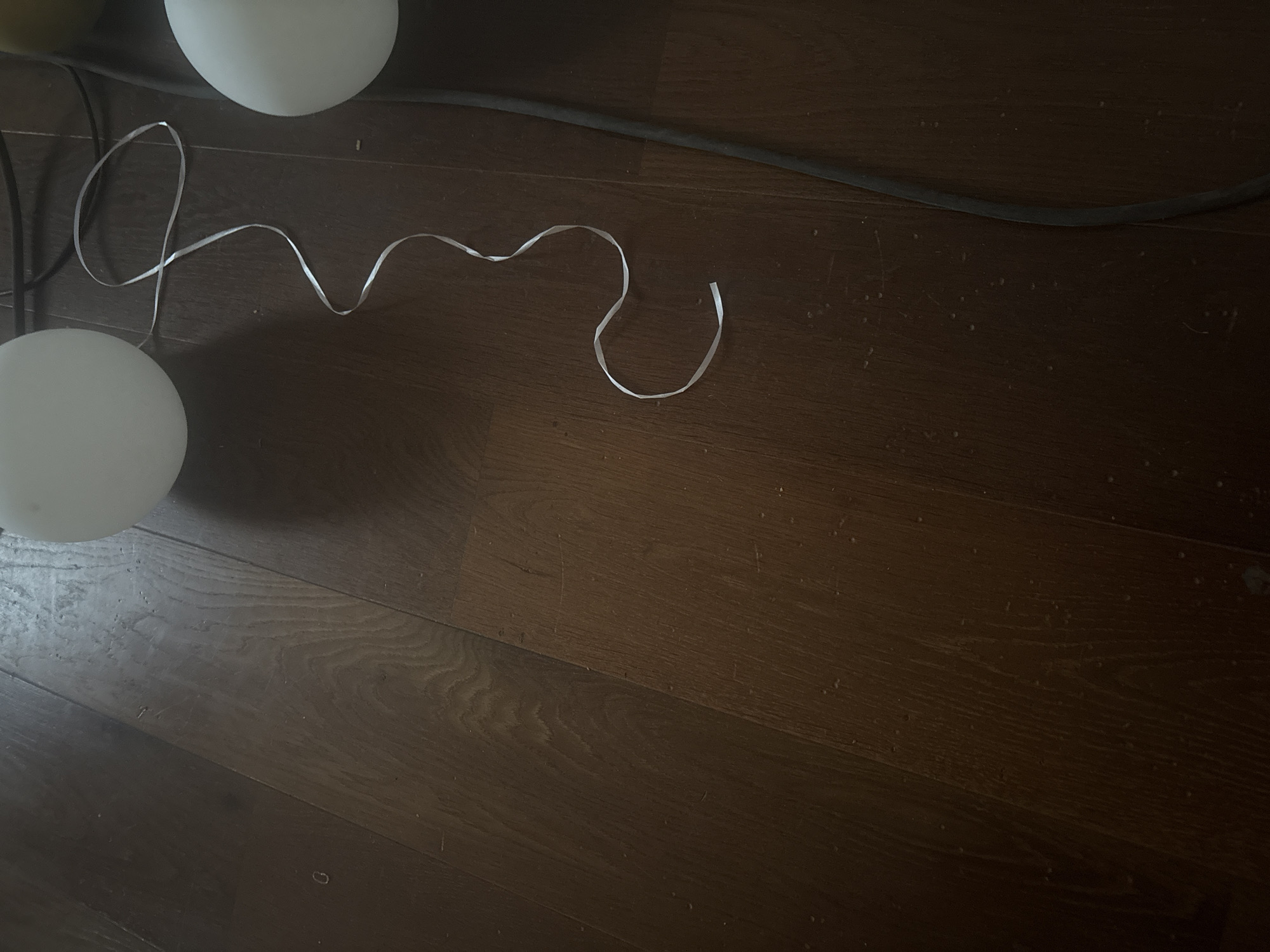The image shows a close-up view of a dark hardwood floor, with a white ribbon and a black cable lying on it. There are also two round, white objects, possibly balloons or lights, partially visible in the upper right portion of the frame. The lighting suggests an indoor setting, possibly a room or a studio. There is no indication of any specific location in the city of Orekhovka, Russia, nor are there any people or interactions depicted. The scene is static and devoid of any narrative context.
