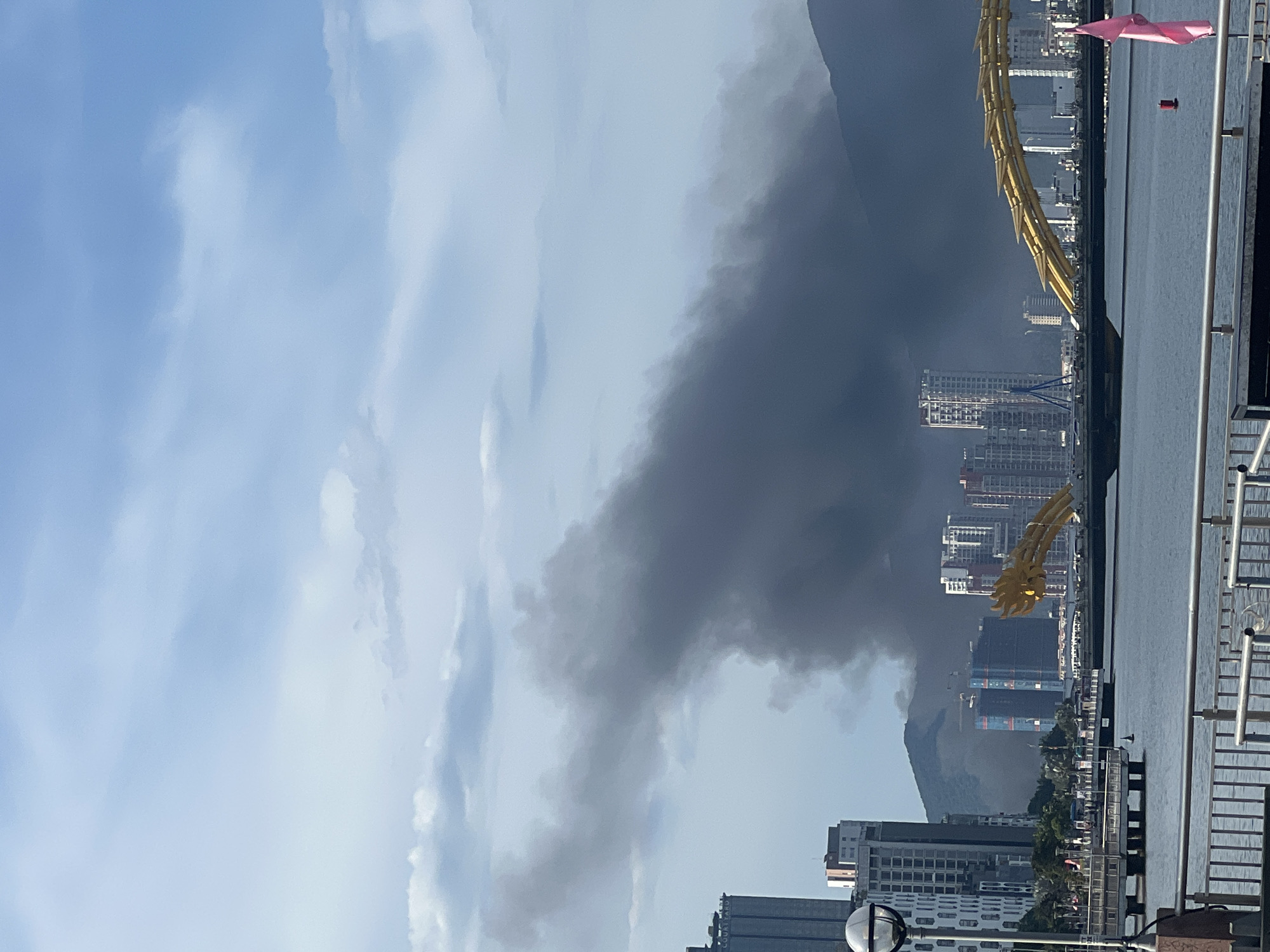 The image shows a view of the city of Đà Nẵng, Vietnam, with the Dragon Bridge prominently visible in the foreground. A large plume of dark smoke is rising from behind the city buildings and mountains in the background, obscuring a portion of the sky. The smoke appears to be a result of a fire or industrial activity. The sky is partly cloudy, with patches of blue visible between the clouds. The Han River flows in the foreground, with a promenade and railings along its bank. Several modern buildings and skyscrapers are visible in the cityscape, along with a bridge under construction. A pink flag is also visible in the bottom right corner of the image.