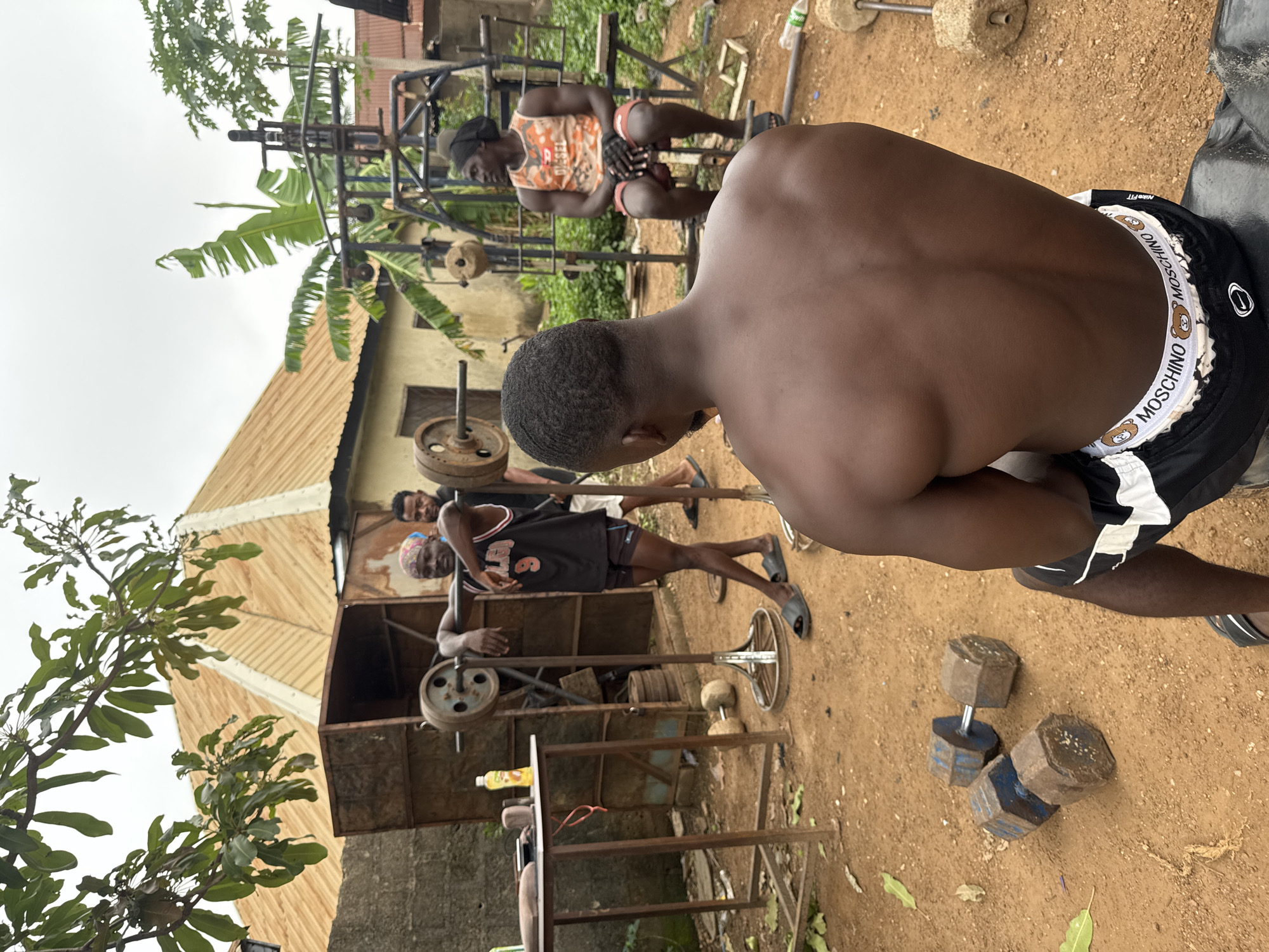 In Akwanga, Nigeria, an outdoor gym is set up in a dusty yard. Three Black men are present. In the foreground, a man from behind, shirtless and wearing black and white "Moschino" branded shorts, is seated on a dark object, possibly a tire. To his left, a rack with a barbell loaded with weights is visible, and a table with a bottle of drink and other items stands nearby. In the middle ground, a man wearing a black basketball jersey with the number 9 and a bandana rests his arm on the barbell rack, while another man in an orange tank top and red shorts sits on a weight bench, wearing black gloves. Behind them, a homemade weight machine and various weights are scattered around. The environment includes a building with a corrugated iron roof, banana trees, and other foliage. The ground is a reddish-brown dirt.