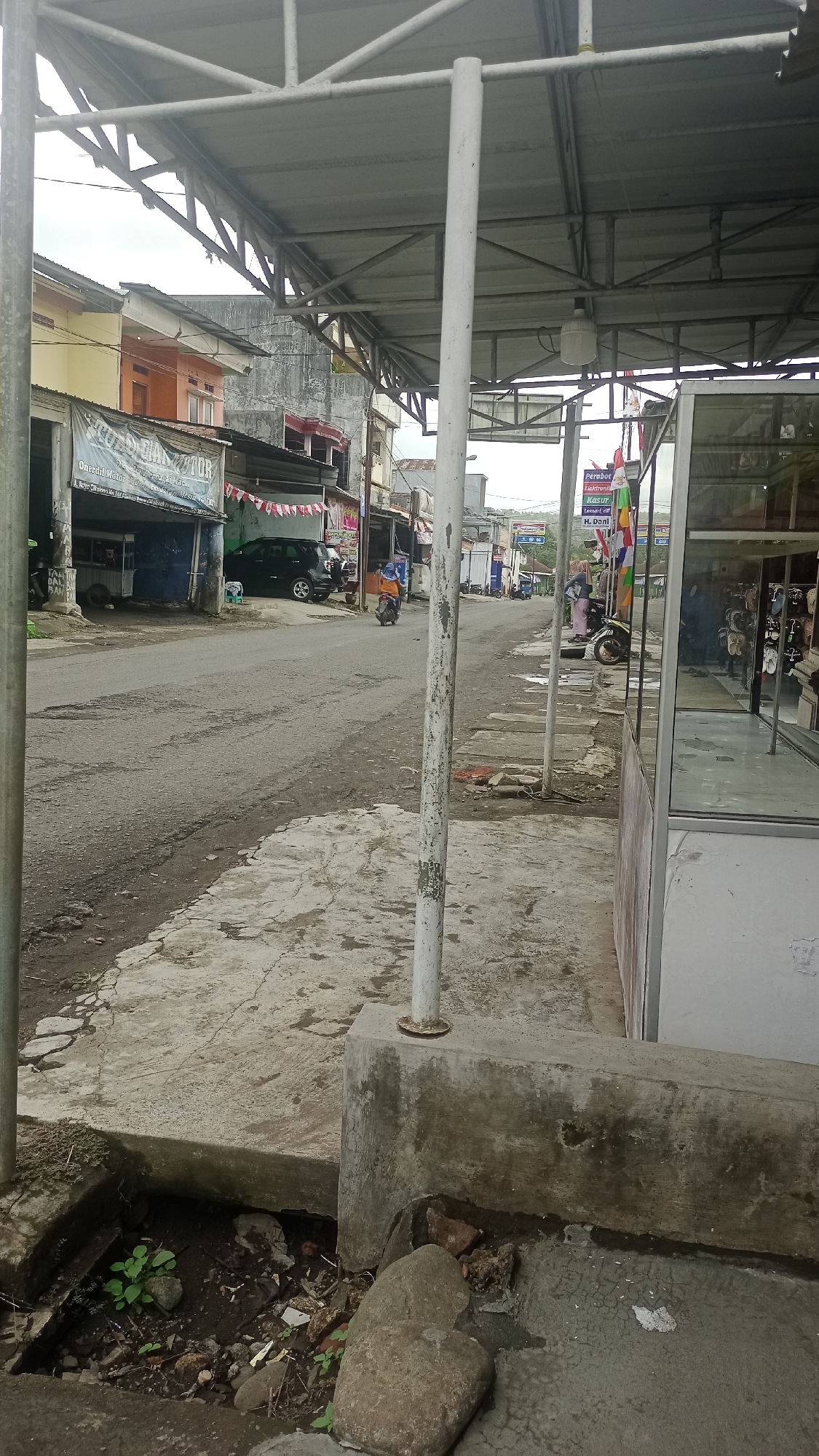 The image depicts a street scene in Cikalong, Indonesia, characterized by a covered walkway and shops lining the road. A grey asphalt road with visible wear and tear runs through the center, with a concrete sidewalk to the right and a section of uneven ground with some vegetation and rocks to the left foreground. Several structures are visible, including shops with open fronts and covered awnings. One shop on the left displays a banner with Indonesian text and what appears to be a logo for a motorcycle repair business. Further down the road, a black SUV is parked, and a person on a motorcycle, wearing an orange shirt and blue pants, is riding away from the viewer.
To the right, a shop with a large glass display window showcases various items, possibly footwear, arranged on shelves. Several colorful flags are displayed outside this shop. The sky above is overcast, suggesting a cloudy day. The overall impression is of a typical, somewhat modest urban street in a region that experiences a tropical climate, as indicated by the open-fronted shops and the presence of greenery. The structures appear to be primarily made of concrete and corrugated metal roofing. The scene is quiet, with limited activity.