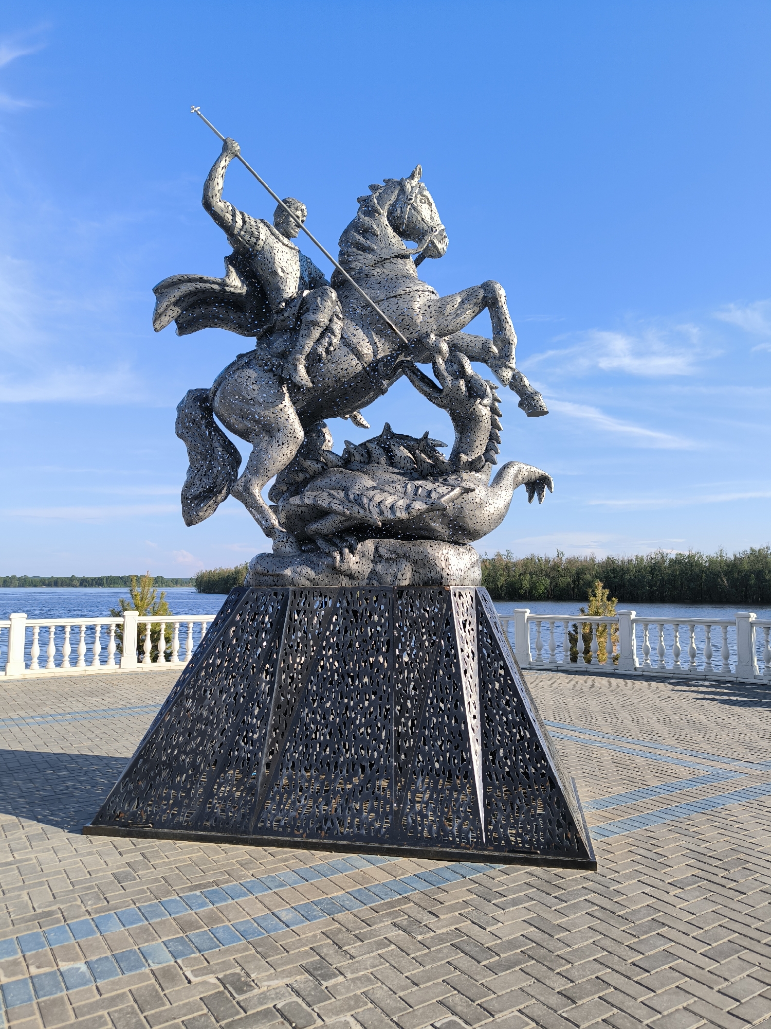 The image shows a metal sculpture of Saint George slaying a dragon, located on a paved plaza next to a body of water. The sculpture is detailed, with the horse rearing and Saint George poised to strike the dragon with a spear. The base of the sculpture is a dark, intricately patterned pyramid. In the background, a white railing lines the waterfront, with trees and a blue sky with wispy clouds visible. This sculpture is located in Surgut, Russia, on the Embankment of the Ob River.