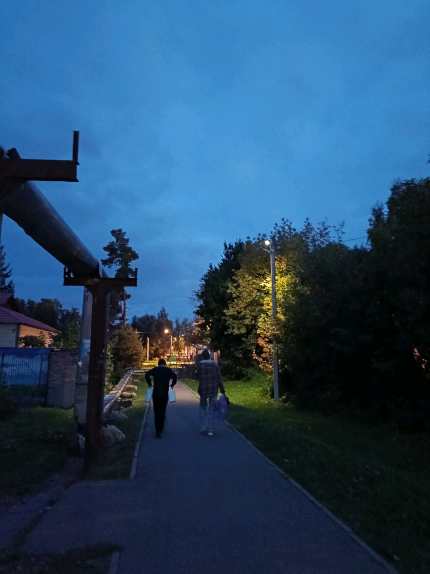 A low-angle, vertical shot captures two individuals walking away from the camera down a paved pathway in Vereya, Russia, during twilight. The sky is a deep blue, transitioning from a brighter hue near the horizon, with scattered clouds. To the left, a large, rusty pipe supported by a metal structure dominates the foreground, obscuring a portion of a residential building with a red roof and a blue fence. Green grass and some foliage are visible on the left side of the pathway, while the right side is bordered by dark trees and a grassy slope. A single street lamp illuminates the path ahead, casting a yellowish glow on the trees. The person in front is wearing a dark long-sleeved shirt and black pants, carrying two white bags. The person behind them wears a plaid shirt and light-colored pants, carrying a purple bag. The overall scene suggests an evening stroll on a suburban street in Vereya.