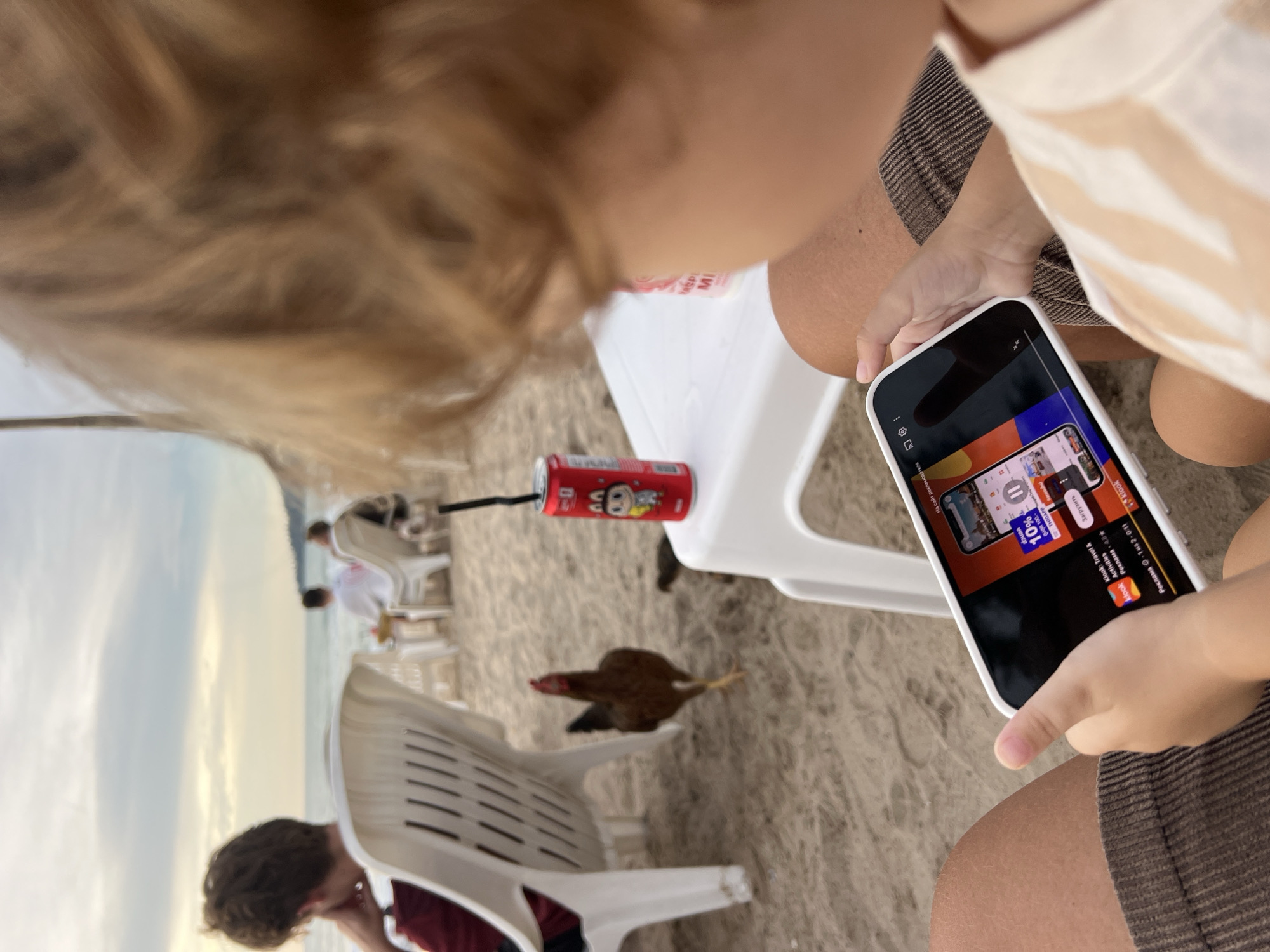 In the city of Choeng Thale, Thailand, at a beachside establishment, a young child in the foreground is focused on a smartphone. The child's face is obscured, but their hands are visible, holding the phone. The phone screen displays an advertisement for "Klook Travel & Activities" offering a 10% discount, with the Russian word "Загрузить" (Download) visible. In the background, out of focus, a person sits in a white plastic chair facing the sea. Further in the background, a few other individuals are seated at tables. A brown chicken walks on the sand near a red can with a cartoon character. The setting is a sandy beach with the ocean and a cloudy sky visible in the distance.