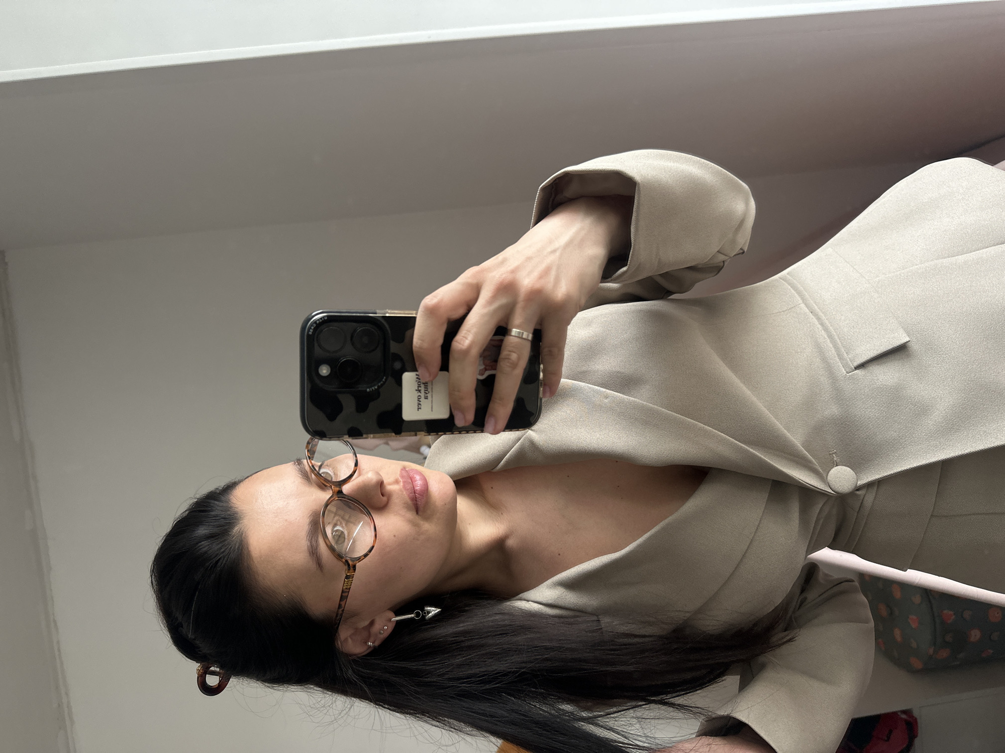 A woman takes a selfie indoors. She has dark hair, glasses, and is wearing a light-colored blazer. She is holding a smartphone with a patterned case. The background appears to be a plain white wall. There is no information given in the images or by the user that confirms the location to be Chaikovsky, Russia.