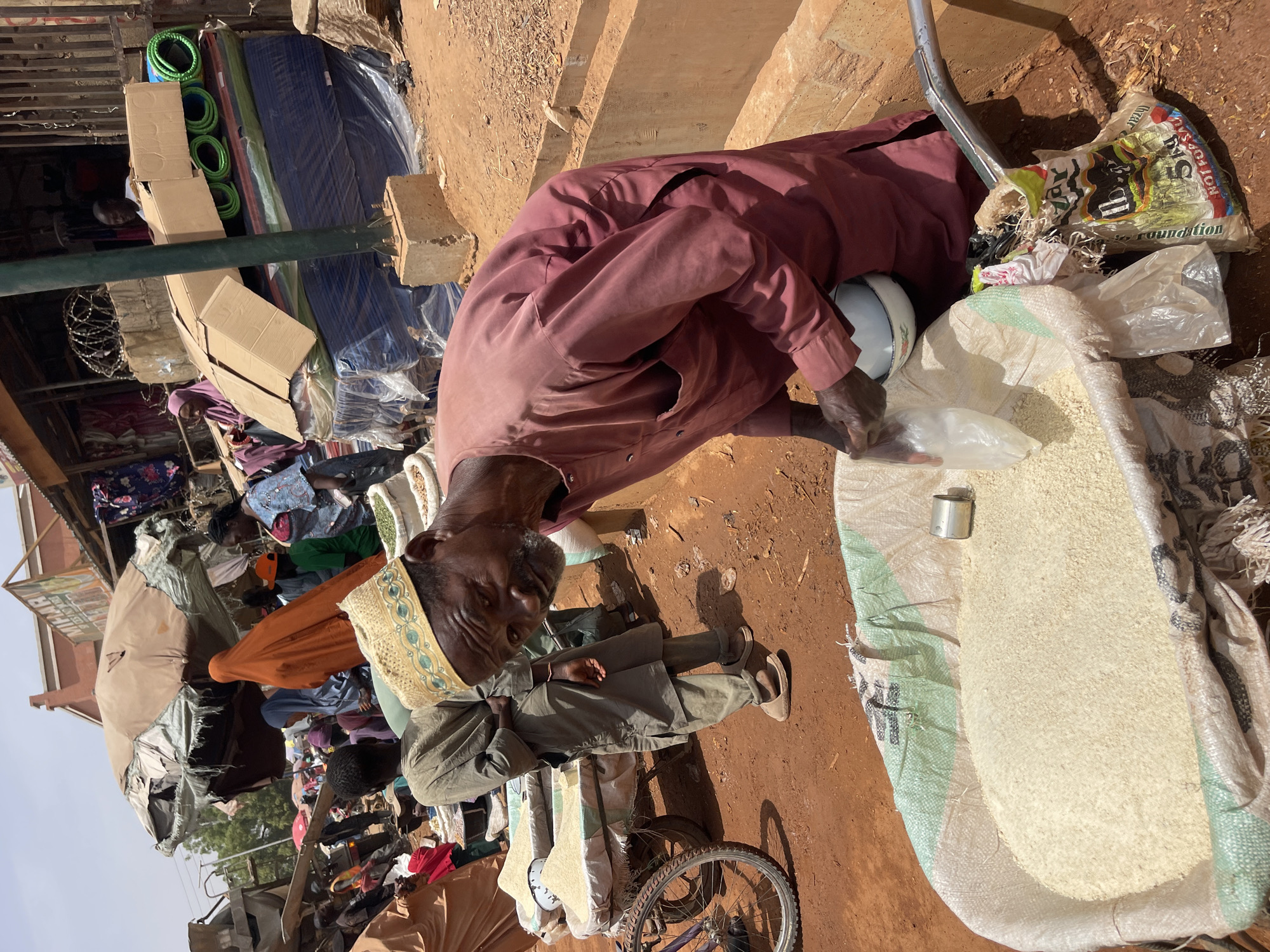 In Zaria, Nigeria, an elderly man wearing a maroon robe is filling a small plastic bag with rice from a large sack. He is using a metal container as a scoop. Another man wearing a green robe and a patterned hat stands nearby. The scene is in an outdoor market area with various goods, including mattresses and household items, visible in the background. A bicycle is partially visible at the bottom of the frame.