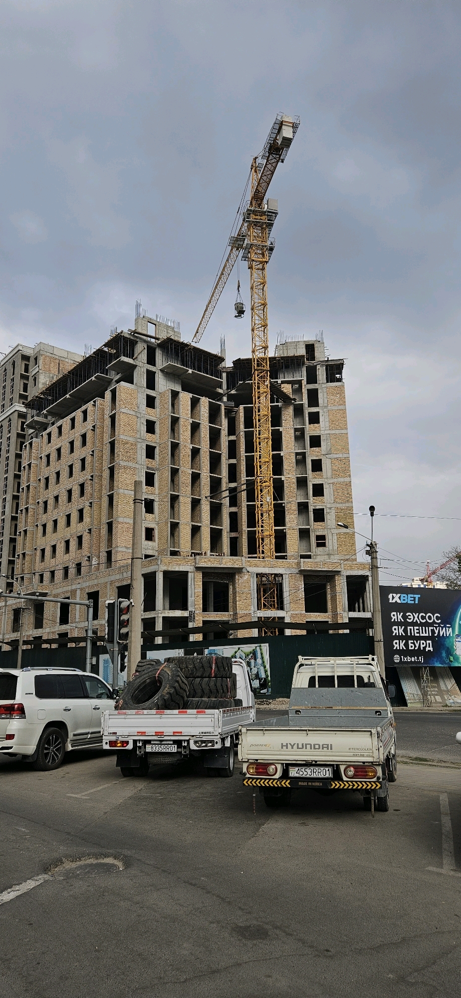 The image depicts a construction site in Dushanbe, Tajikistan, with a large unfinished building and a yellow construction crane dominating the scene. The building is made of brick and concrete, with many windows and exposed rebar. In the foreground, two pickup trucks are parked on the street. The truck on the left is a white Toyota Land Cruiser. The truck in the center is a white Hyundai Porter carrying a stack of large tires. The truck on the right is also a white Hyundai Porter. A billboard advertising "1XBET" is visible in the background. The sky is overcast.