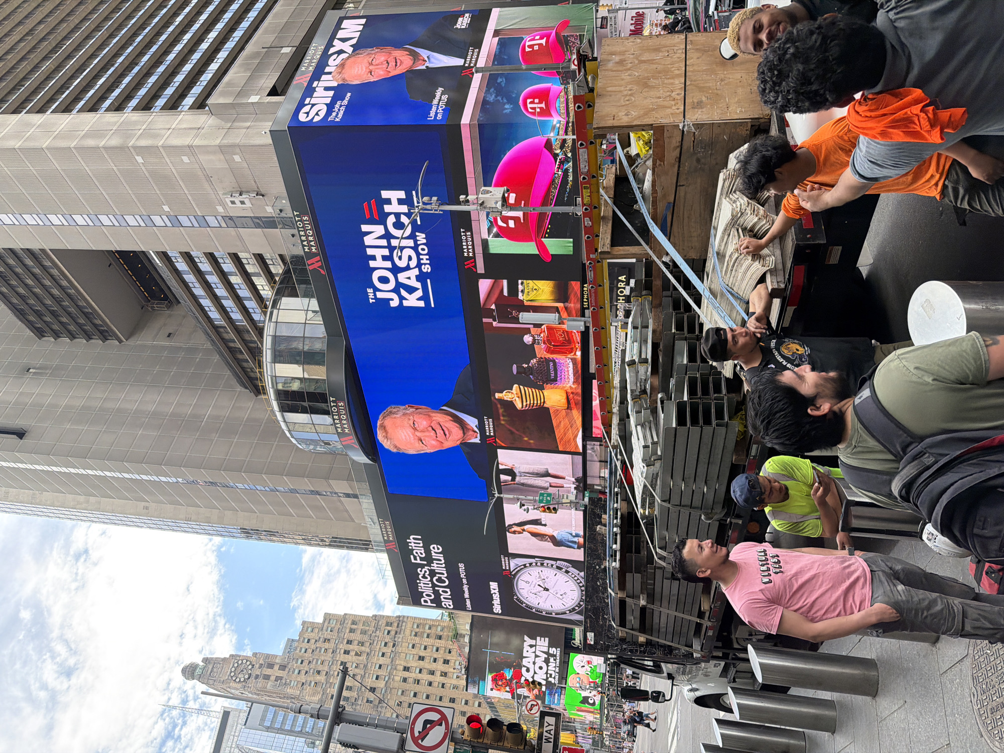 The image shows a street scene in New York City, United States. A group of men are on the sidewalk in front of a building with large LED billboards. The billboards feature advertising for "The John Kasich Show" on SiriusXM, T-Mobile, and perfume products. The billboards also display the Marriott Marquis logo.
A traffic light and street signs are visible in the lower part of the image. In the background, there is a tall building with a clock tower. There are several people on the sidewalk.