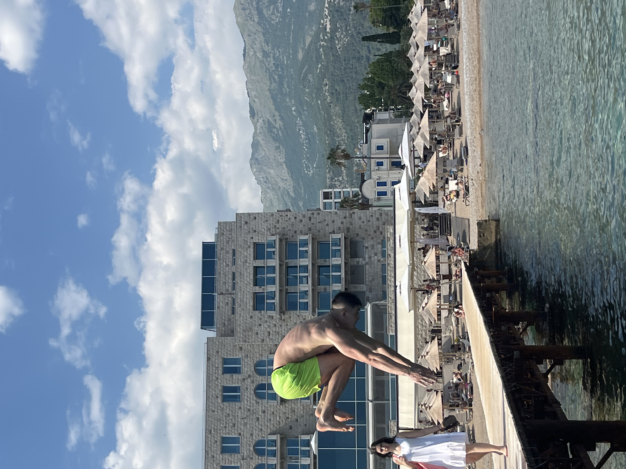 A man in neon green shorts is captured mid-jump, poised to dive from a pier. He is athletic, with a lean build, and his body is taut with anticipation. His arms are extended forward, fingers splayed, as if preparing to slice through the water. Below him, a woman in a white dress stands on the pier, her gaze directed towards him. Her presence suggests she is either waiting for him or observing his dive.
The scene is set on a pier overlooking a clear, turquoise body of water. In the background, a bustling beach is visible, lined with rows of beige sun umbrellas and lounge chairs, many occupied by sunbathers. Behind the beach, a modern, multi-story building with a facade of stone and large blue-tinted windows rises. Further in the distance, imposing, verdant mountains dominate the skyline, their slopes textured with shadows and vegetation.
The sky is a vibrant blue, dotted with fluffy white clouds. The sunlight suggests it is daytime, likely a warm and pleasant day, ideal for a beach outing. The overall impression is one of a holiday atmosphere, with leisure and recreation at the forefront. The focus is on the dynamic action of the diver, contrasted with the relaxed scene of the beach and the grand natural backdrop. There is no visible text in the image.