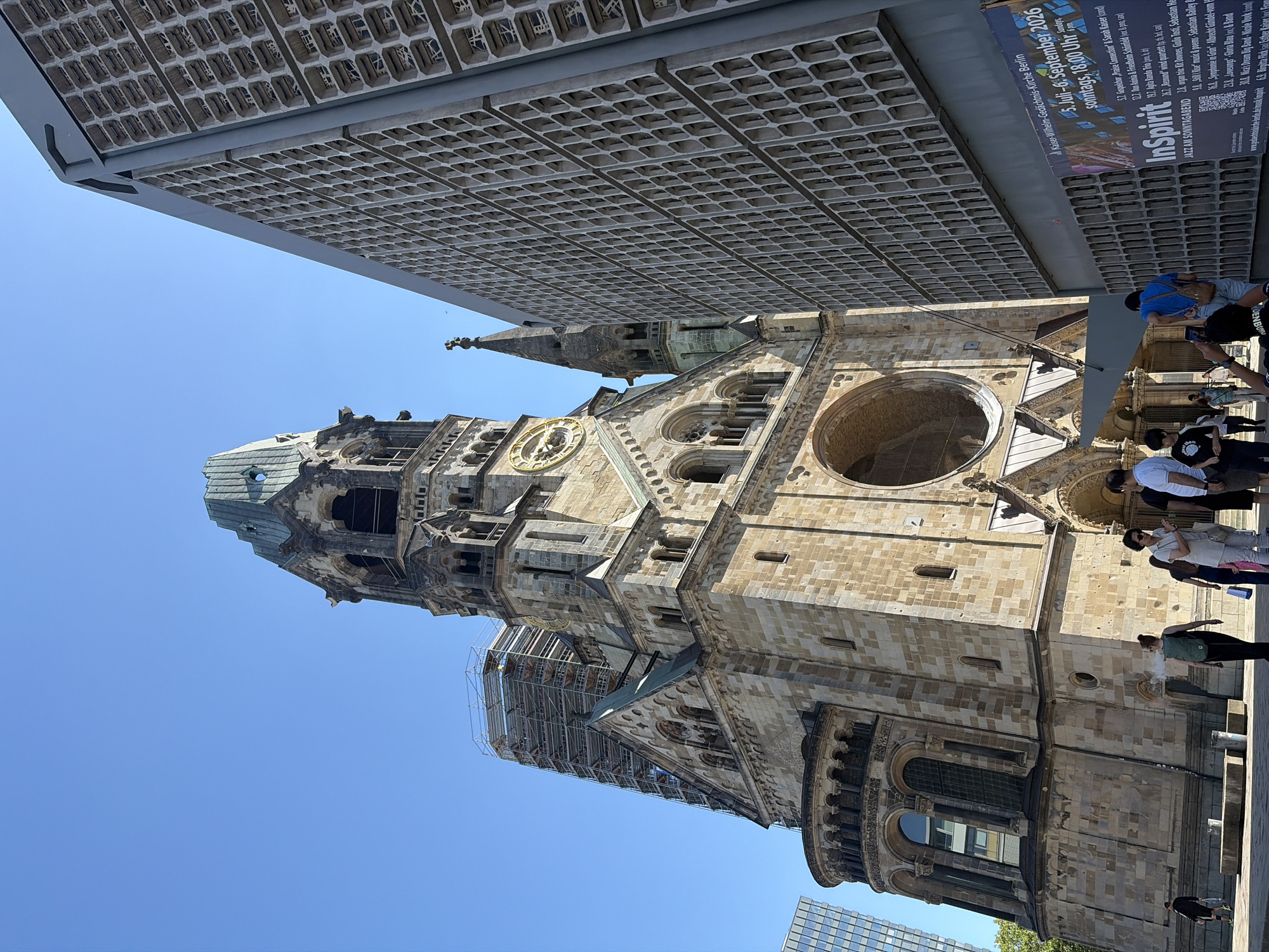 The image shows the Kaiser Wilhelm Memorial Church in Berlin, Germany. The church's ruined tower, a prominent landmark, is on the left, partially covered in scaffolding. To its right is a modern, hexagonal-patterned building. Several people are visible in the foreground, some walking and others taking photos. A poster advertising an event called "InSpirit" is affixed to the modern building. The sky is clear and blue, indicating a sunny day.