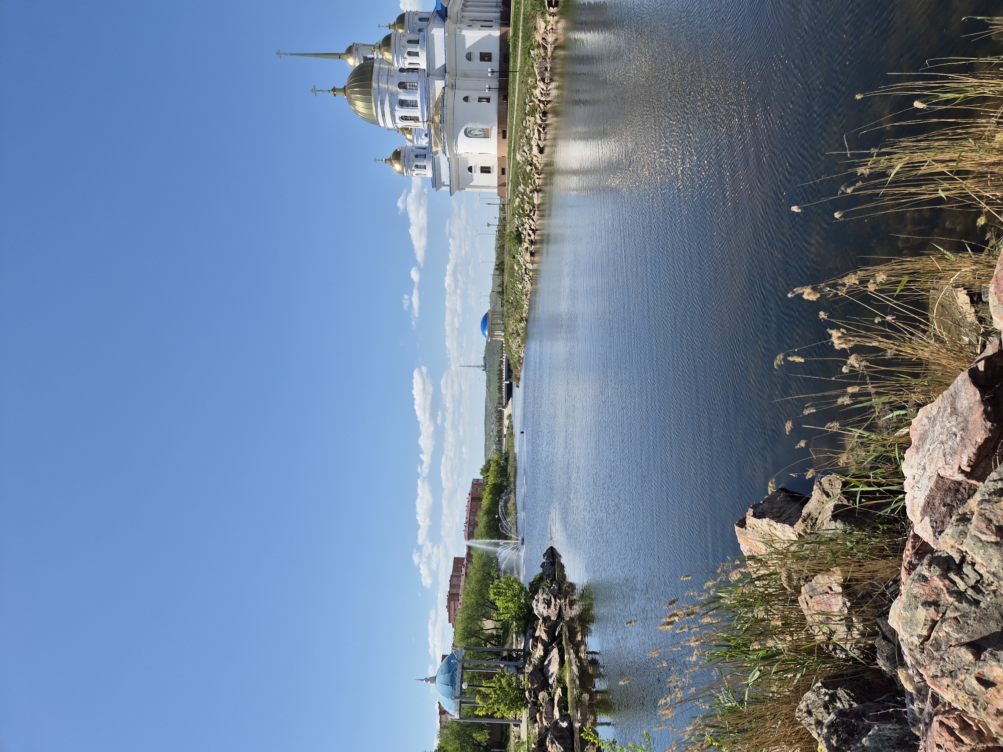 The image depicts a cityscape in Kokshetau, Kazakhstan, with a prominent body of water extending through the scene. On the upper right of the image is the Holy Resurrection Cathedral, recognizable by its white facade and multiple gold domes. The lake reflects the sky and the cathedral's architecture. On the lower-left, there are trees, landscaping, and a gazebo with a blue dome. A fountain is visible in the distance. In the lower right, there are rocks and reeds along the water’s edge. The sky is blue and cloudless.