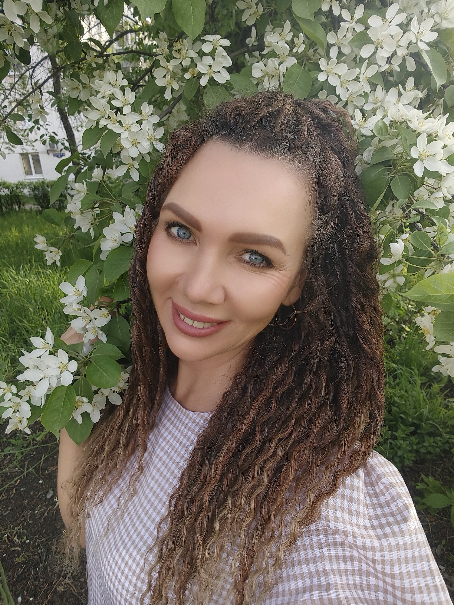 A woman with blue eyes and brown curly hair stands in front of a flowering tree with white blossoms. She is wearing a light brown and white checkered dress and gold hoop earrings. Her hair is styled with a portion pulled back on top of her head. Behind her, there is a building with windows and green grass and foliage, likely in the city of Unknown, Unknown.