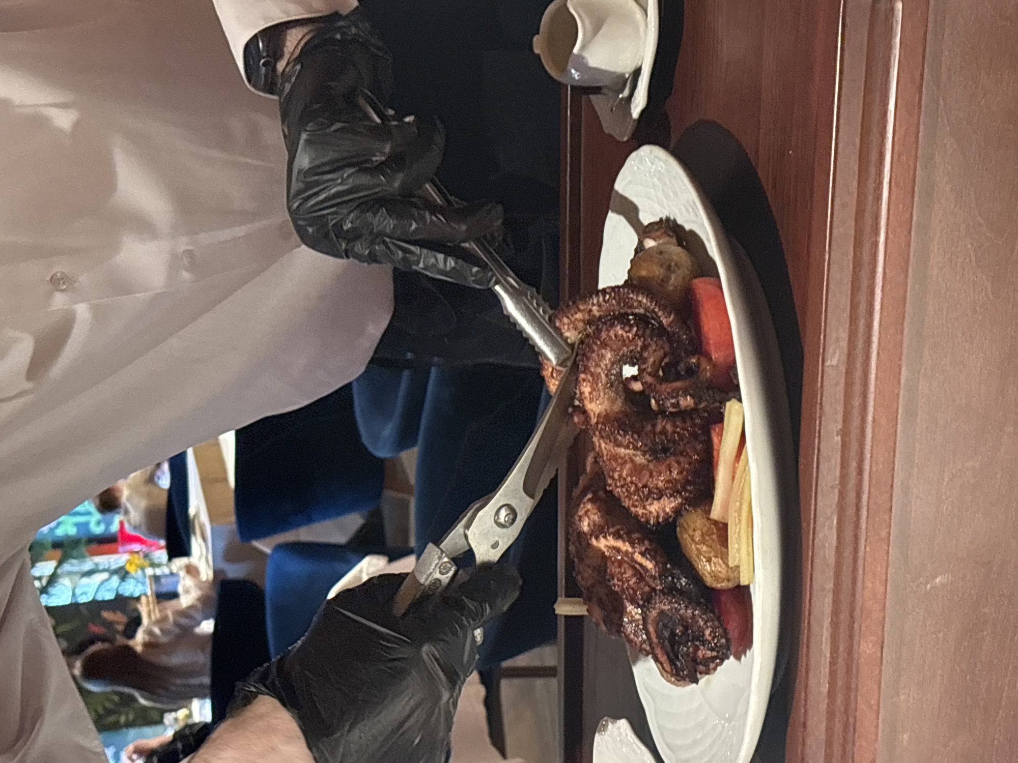 In a restaurant located in Saint Petersburg, Russia, a server wearing a white long-sleeved shirt, dark trousers, and black disposable gloves is actively portioning an octopus dish on a dark brown wooden surface. The server's left hand holds a pair of metal kitchen shears, while their right hand grips a piece of food with metal tongs, positioned over a white, fluted plate. The plate contains several dark, textured pieces of grilled or roasted octopus, accompanied by red sliced vegetables (possibly tomatoes) and yellow-beige elongated vegetables (possibly roasted potatoes or pickles). To the right of the plate, a small white creamer or gravy boat rests on a matching saucer with a spoon. The background reveals a softly lit dining area with multiple patrons seated at tables, one woman with long hair visible from behind. The far background features large windows or decorative panels with intricate, colorful glass designs in shades of blue, green, and red, creating a distinct visual element. Blue upholstered chairs are partially visible at the dining tables.