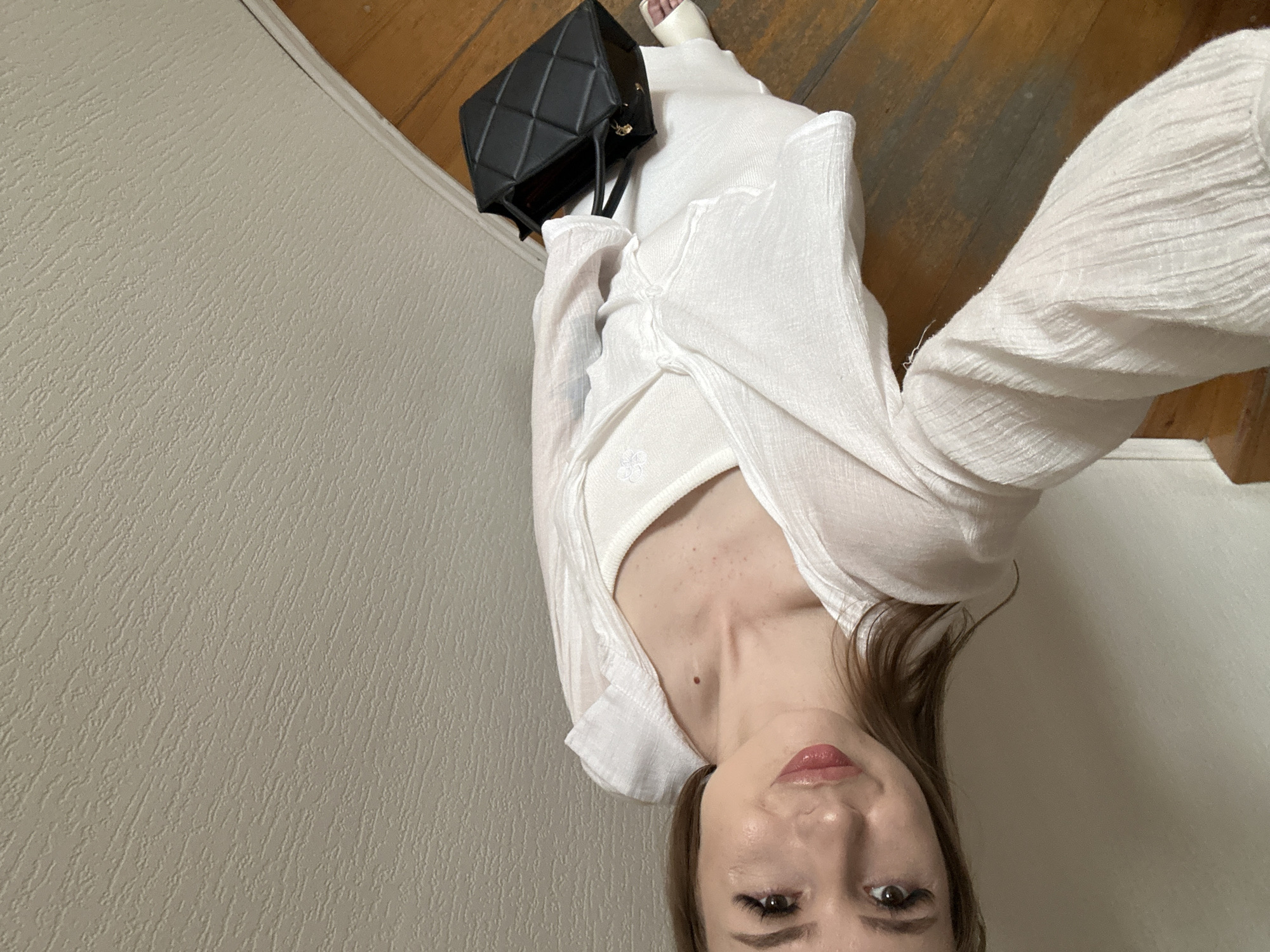A woman stands on a wooden staircase, holding a black purse. She is wearing a white tank top and a white linen dress with a white linen shirt over it. The walls in the background are textured and light-colored. The floor is made of polished wood planks. The woman's hair is brown and her makeup is subtle. She is looking at the camera with a slight smile.