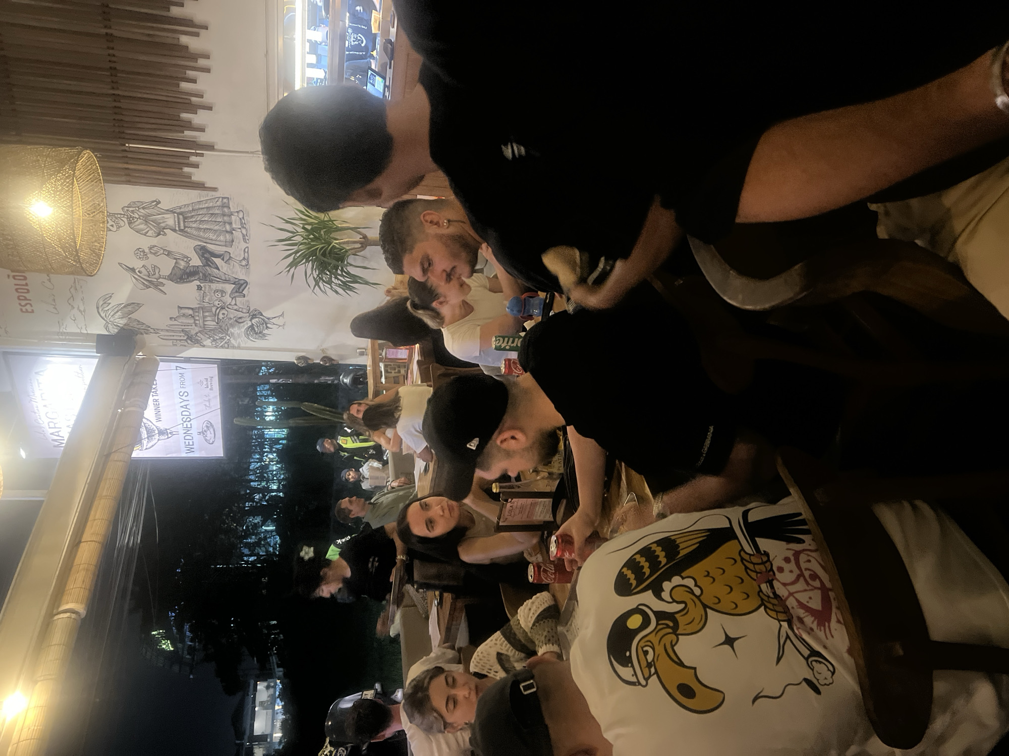 The image depicts an indoor scene at a restaurant or bar in North Kuta, Indonesia, during the nighttime. A group of individuals, primarily young adults, are seated around tables, engaged in conversations and consuming food and drinks. The lighting is dim, with artificial lights illuminating the space, creating a warm and casual ambiance.
In the foreground, a person wearing a white t-shirt with a cartoon bird graphic is visible. They are seated, and their arm is resting on a wooden chair. To their right, another person wearing a black t-shirt is partially visible, their arm and shoulder in focus.
Further back, several other people are seated at tables. One woman with long dark hair is looking to her left, wearing a light-colored top. Beside her, a woman with blonde hair is wearing a striped sweater and looking down at the table. A man with a short beard and dark hair is looking towards the right side of the frame, wearing a black t-shirt. Another man with short dark hair and a light-colored shirt is visible behind him.
The restaurant's interior features wooden elements, including ceiling beams and wall paneling. Decorative murals adorn the walls, one of which appears to depict skeleton figures in a festive or cultural scene. There are also signs with text, one prominently displaying "MARGARITA." In the background, glimpses of other patrons and restaurant staff can be seen, along with menu items and what appears to be a bar area with equipment. Outdoor foliage and trees are visible through a large open doorway or window, suggesting the restaurant might be partially open-air or has an adjacent outdoor space. The overall impression is that of a lively, social gathering in a tropical setting.