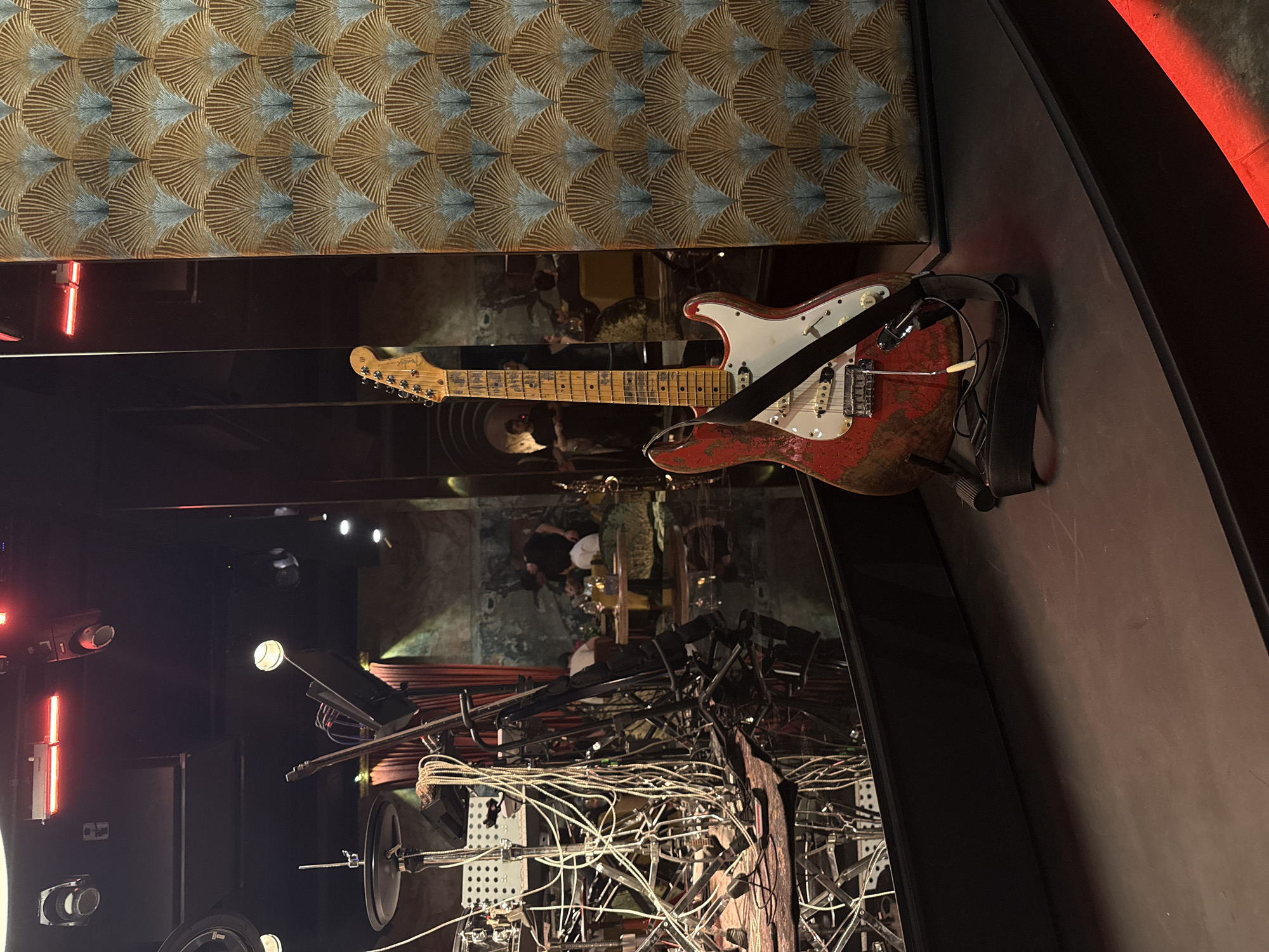 An electric guitar, appearing to be a heavily worn red and white Fender Stratocaster, lies prominently in the foreground of an indoor scene. The guitar exhibits significant wear and tear, with much of its red paint chipped away, revealing a white or lighter undercoat, particularly on its body. A black strap is attached to it, and the "Fender" logo is visible on its headstock. The guitar rests on a sleek, dark, reflective surface that appears to be part of a stage or display area.
Above the guitar, a wall or ceiling is covered with a distinctive patterned material featuring repeated fan-like or shell-like motifs in shades of gold, brown, and muted blue or grey. To the left and in the deeper background, complex metallic structures intertwined with numerous white cables and wires are visible, suggesting an elaborate sound or lighting system, or perhaps a modern art installation. Various spotlights and red neon light strips illuminate parts of this intricate setup.
Reflected in the dark surface behind the guitar, and possibly through a transparent partition, are several people, suggesting the location is a lively venue like a bar, restaurant, or club. The artificial lighting, including the red neon elements, indicates an evening or nighttime setting. The overall atmosphere is modern and industrial, with a strong musical or performance arts undertone, reinforced by the presence of the guitar and the technical infrastructure. The location context indicates this is in Yerevan, Armenia.