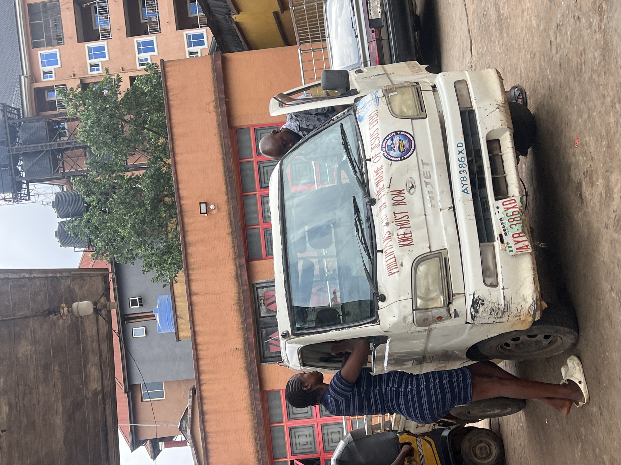 In the city of Nkpor, Nigeria, on a street with an overcast sky, a white Daihatsu Hi-Jet truck is parked. The truck's front bumper is dented and scratched. On the windshield, there is text that reads "PHILIPPIANS 2:10 IN THE NAME OF JESUS EVERY KNEE MUST BOW". Below this text, a circular sticker with a logo and the words "ASSOCIATION OF..." is visible. A license plate with the registration "AYB 386XD" is affixed to the front of the truck. A young woman with braided hair, wearing a blue and white striped dress and white Crocs, stands by the driver's side door, leaning in. A man with a shaved head, wearing a patterned shirt, is leaning into the truck from the passenger side door, which is open. To the left of the truck, a portion of a yellow and black auto-rickshaw is visible. In the background, there are multi-story buildings with orange and gray facades and red-trimmed windows, as well as trees and water tanks on a metal structure.