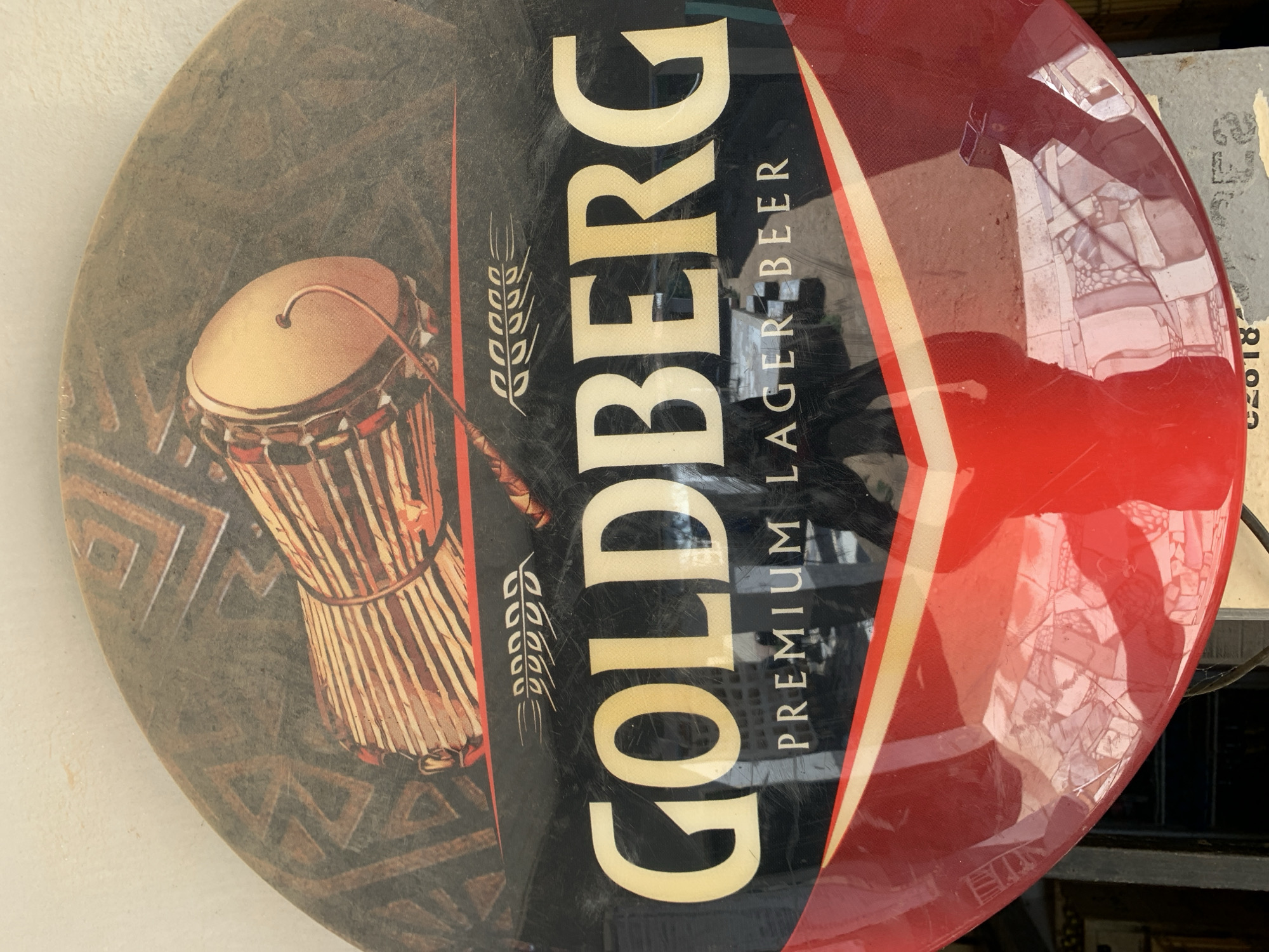 The image shows a circular Goldberg Premium Lager Beer advertisement sign. The sign is divided into three sections. The first section, on the left, features an image of a djembe drum against a patterned, somewhat weathered background. The middle section is black and displays the brand name "GOLDBERG" in large, bold, off-white lettering vertically. Below the brand name, it reads "PREMIUM LAGER BEER" in smaller, vertically aligned lettering. Wheat stalks flank the left side of the lettering. The third section, on the right, is predominantly red with a yellow and white stripe creating a visual break. Reflections are visible on the shiny red surface of this section. The sign appears to be aged, with visible wear and discoloration. The location in the city of New Stadium, Nigeria is not explicit in the image, however Goldberg is a Nigerian Lager.