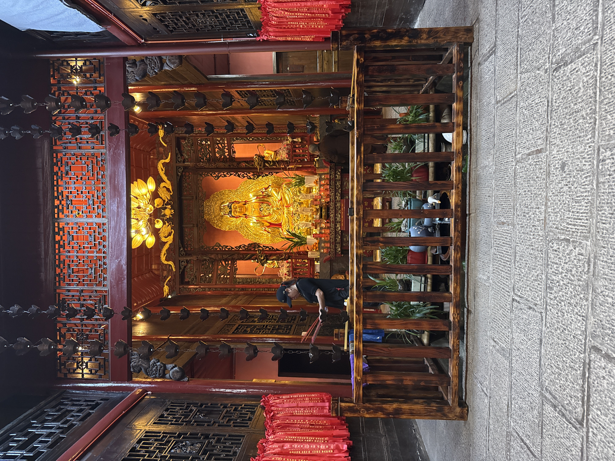 The image captures a scene at Furong Ancient Town in Xiangxi Tujia and Miao Autonomous Prefecture, China. A golden Buddha statue is prominently displayed in an ornate, red and gold-toned interior, embellished with intricate carvings and decorative lamps. A man wearing a black shirt and cap is bending over, holding incense sticks, possibly in prayer. A wooden structure, resembling a balcony, stands adjacent to the temple-like building, adorned with potted plants and rows of small metal ornaments hanging from the ceiling. Stacks of red fabric, possibly prayer cloths, are visible near the bottom of the frame. The exterior is paved with grey stone.