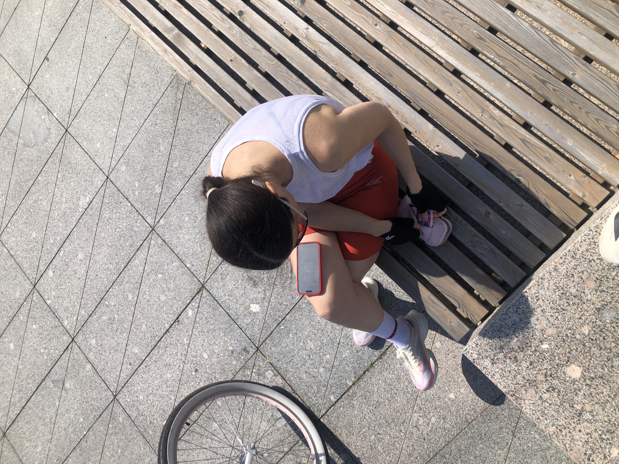This is a high-angle, medium shot of a woman sitting on a wooden bench outdoors, tying her shoelaces. She has dark hair pulled back in a ponytail and is wearing a white tank top, red shorts, white socks with pink stripes, and pink and white athletic shoes. She is also wearing white earbuds and has a red smartphone attached to her left thigh with a phone holder. A bicycle wheel is visible in the lower left corner of the frame. The ground is paved with large grey tiles arranged in a diagonal pattern. The bench is made of wooden slats. The scene appears to be in a public outdoor space, possibly a park or promenade, given the bench and paving. The lighting suggests it is daytime, with a bright sun casting shadows. The overall mood is relaxed and casual, suggesting the woman is preparing for or has just finished an activity like cycling or running. The presence of the bicycle and athletic wear indicates an active lifestyle. No visible text is present in the image.