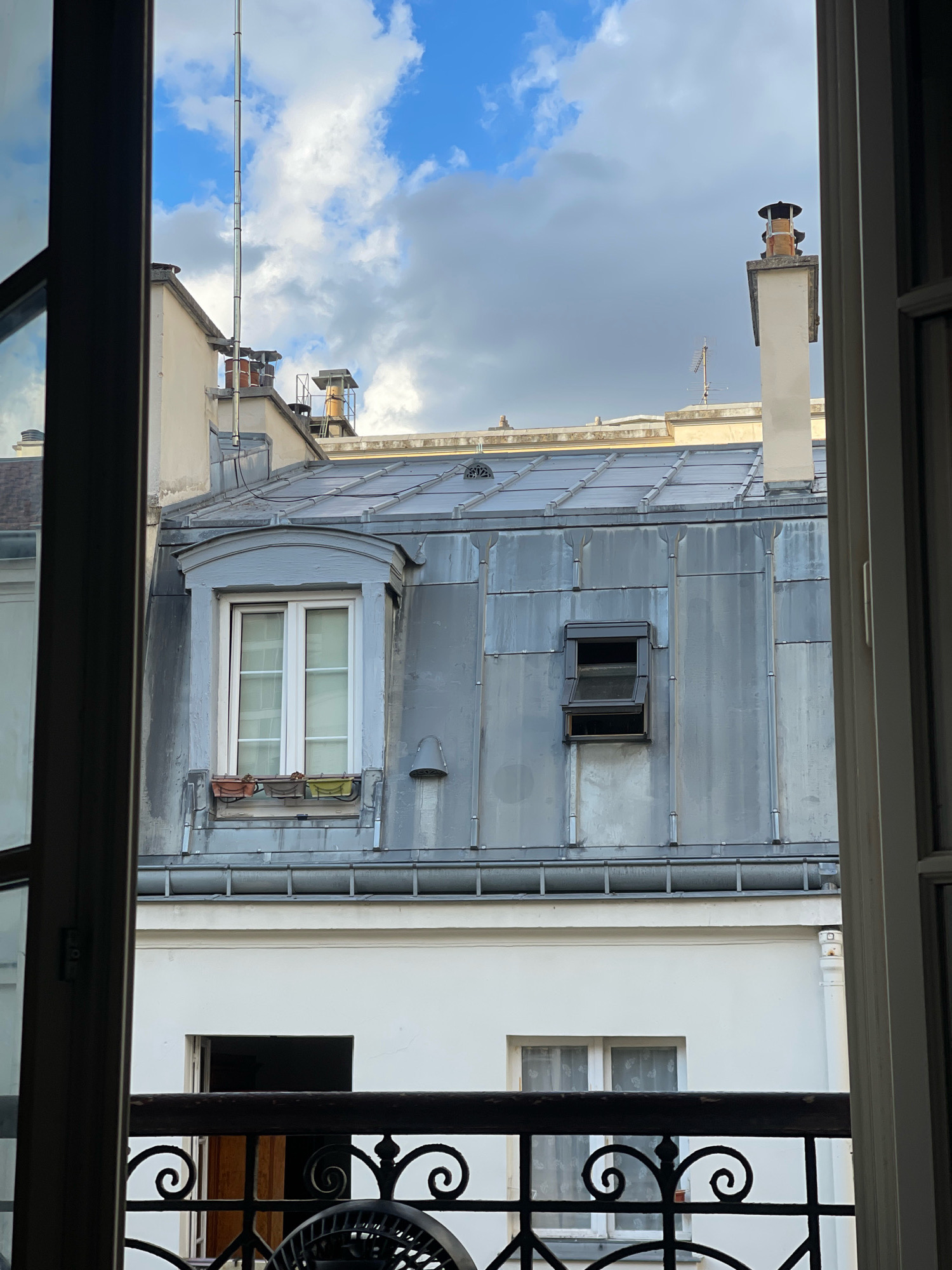 The image shows a view from a window in Paris, France, looking out at rooftops and the sky. The foreground features the dark interior of a room, with the edges of a window frame visible on the left and right. A wrought-iron railing with ornate scrollwork is present at the bottom of the frame, with a portion of a black fan visible below it.
Beyond the railing, the scene opens up to reveal the characteristic zinc rooftops of Parisian buildings. A large, mansard-style roof dominates the center, covered in vertical zinc panels. A dormer window with a gray frame and white-paned glass is centrally located on this roof. Below the dormer window, a narrow planter box holds three small plants. To the right of the dormer, a smaller, dark-framed opening window is visible, partially ajar.
Further back, other rooftops and building structures can be seen. A tall, rectangular chimney with a decorative cap stands on the right side of the frame. Another chimney, made of brick, is visible to the left of it, partially obscured by a taller structure. An antenna, consisting of a series of metallic poles, extends upwards into the sky.
The sky above is a mix of blue and gray clouds, suggesting a dynamic weather pattern. The buildings appear to be made of stone and painted stucco, typical of Parisian architecture. The overall impression is a classic urban landscape in Paris, viewed from a residential window.