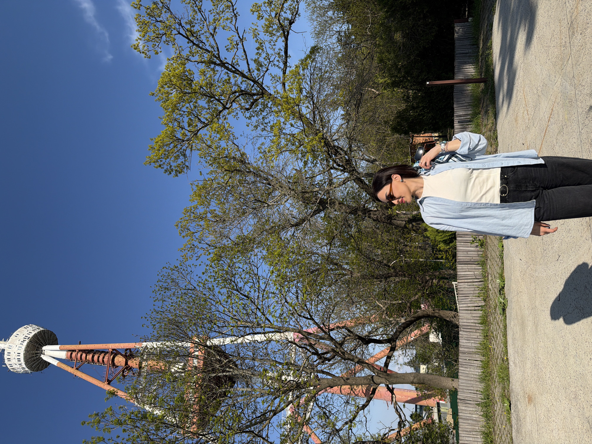 A bright outdoor portrait-oriented image shows a woman standing on a paved path, framed by trees and a communication tower, under a clear blue sky. The woman, positioned on the right side of the frame, is wearing a light blue long-sleeved button-up shirt over a white top, dark pants, and sunglasses. Her dark hair is pulled back, and she is looking downwards and slightly to her right.
To the left and in the background, numerous trees fill the scene. Many of these trees are in various stages of foliage, with some showing fresh, bright green leaves, indicating springtime. Beyond the trees, on the far left, a distinctive white and reddish-orange communication tower with a circular observation deck at its peak is visible, identifiable as the Tbilisi TV Tower.
The ground consists of a light-colored paved path, likely concrete or asphalt, with some patches of grass and a low wooden fence lining the path further back. The day is sunny with a clear, deep blue sky dominating the upper left portion of the image, featuring only a few wispy white clouds. Strong sunlight casts distinct shadows from the trees and the woman onto the path, suggesting a midday or early afternoon setting. No visible text is present in the image.