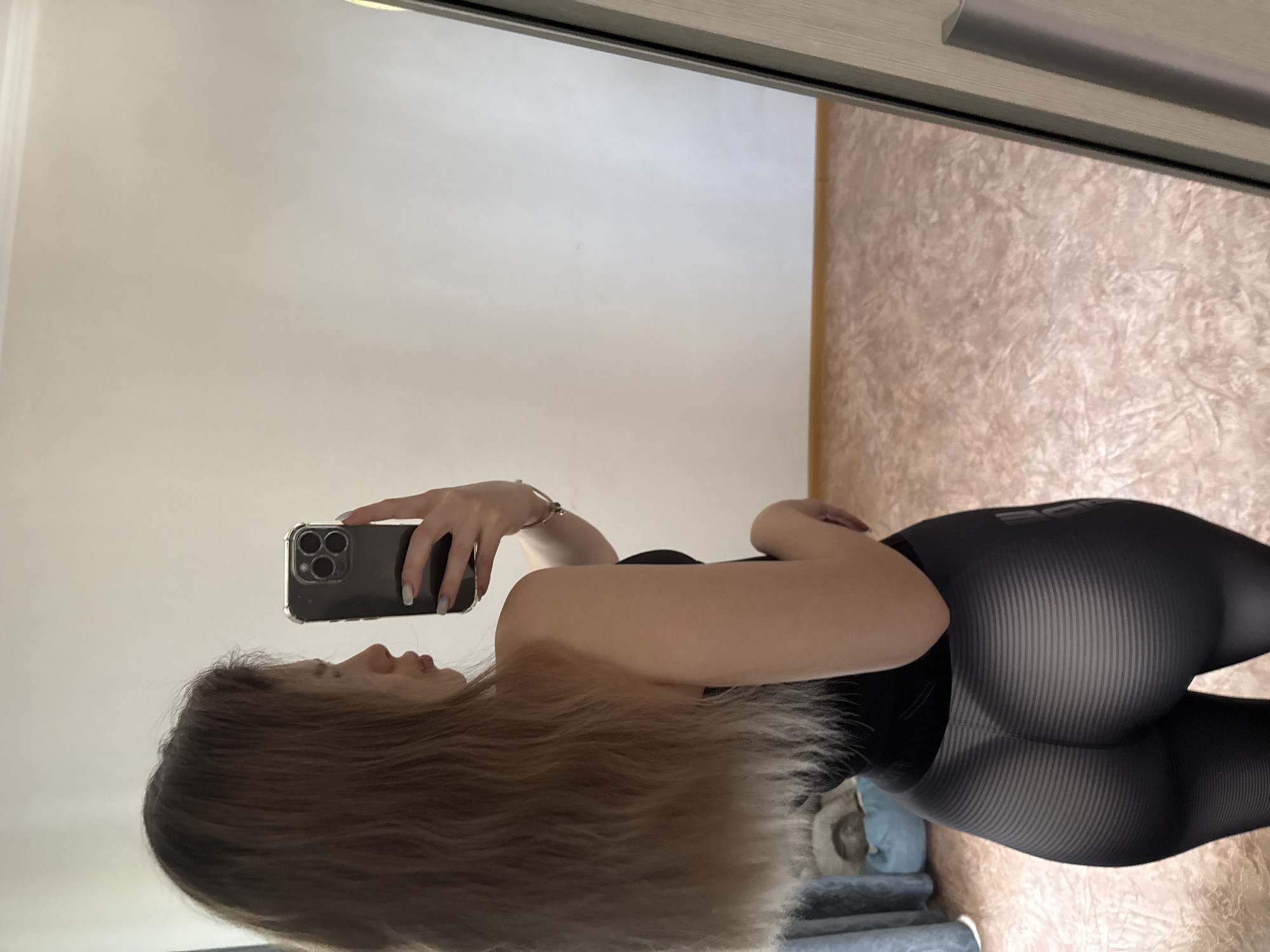 The image, rotated 90 degrees clockwise, captures a person taking a mirror selfie indoors. The subject is a female with long, light brown hair, seen in profile looking towards the phone. She is holding a dark-colored smartphone with a clear case and three prominent camera lenses in her right hand, positioned to capture her reflection in the mirror. She is wearing a dark, possibly black, sleeveless top and form-fitting dark leggings. The leggings feature a subtle striped or ribbed pattern and have part of a word, "BOD," visible on the back of the waistband, likely part of a brand name or the word "BODY." Her nails are manicured with a light polish.
The background visible in the mirror reflection includes a plain light-colored wall on the left and a light brown or peach-colored textured floor or carpet on the right. In the bottom left corner of the reflection, what appears to be part of a bed or furniture with a light blue cushion or blanket is visible. The top edge of the mirror shows part of a wooden frame with a metal handle, suggesting it might be a sliding wardrobe door or a large standing mirror. The lighting is soft and even, typical of indoor conditions. No specific time of day or outdoor environment cues are present.