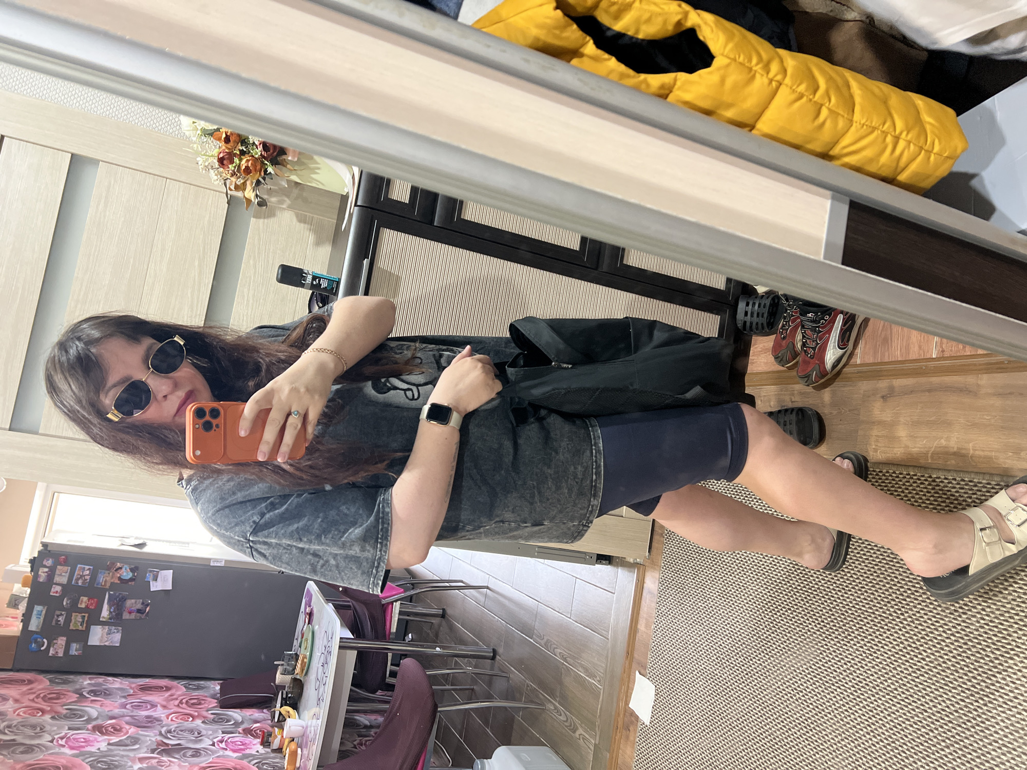 A woman wearing sunglasses is holding an orange smartphone up and taking a picture of herself in a mirror. She is wearing a dark-colored t-shirt, shorts, a gold-colored bracelet, and a smartwatch. Her brown hair is long and loose. In the reflection of the mirror, there is a kitchen with a floral-patterned tablecloth and a gray refrigerator covered in magnets. A yellow puffer jacket, and various pairs of shoes can also be seen in the reflection. The location could be Noviy, Russia.