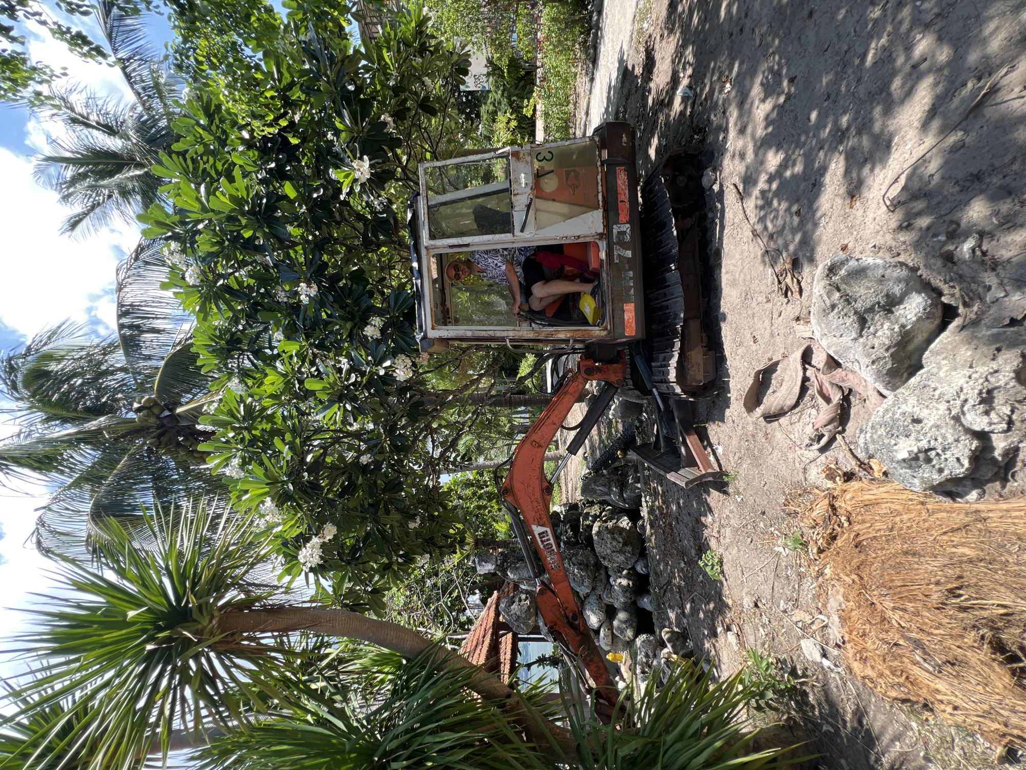 In a sun-drenched outdoor scene in Bac Nha Trang Ward, Vietnam, a man is seated in the cab of an orange and white excavator. The excavator is positioned on a sandy, debris-strewn area with a pile of large rocks to its left. The man, wearing sunglasses and a patterned shirt, is holding the excavator's controls. Lush green foliage, including palm trees and flowering bushes, surrounds the scene. A small, open-air structure with a red tile roof is visible in the background, as is a body of water. The ground is covered in dirt, small stones, and fallen leaves. The excavator's tracks are visible, resting on the ground.