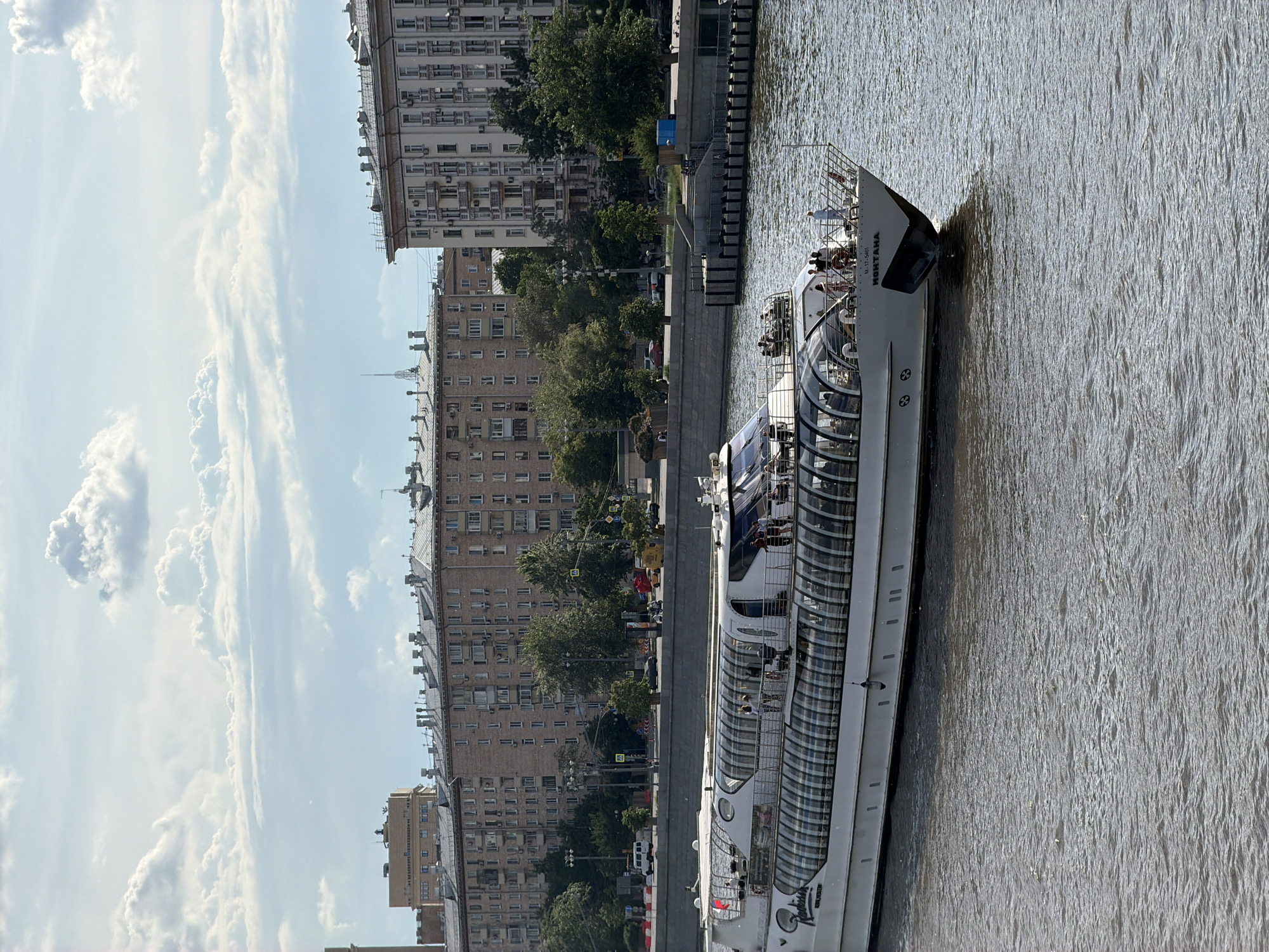This image captures a vibrant daytime scene along the Moscow River in Russia, featuring a modern passenger boat sailing on the water. The boat, prominently named "МОНТАНА" (MONTANA) with a registration number "М-17-5461" visible on its bow, boasts a distinctive glass-enclosed upper deck designed for sightseeing. Several passengers can be seen on both the open-air and enclosed sections of the upper deck, suggesting a leisure cruise.
The river exhibits a gently rippled surface, reflecting the bright sunlight. Along the embankment, a row of green trees provides a natural buffer against the urban backdrop. Behind these trees, multiple large, multi-story buildings line the street, characterized by numerous windows and some external air conditioning units, typical of Moscow's residential and commercial architecture. Vehicles are visible on the road running parallel to the river.
The sky is partly cloudy, with scattered white clouds against a bright blue background, indicating fair and pleasant weather. The overall atmosphere is one of a calm and enjoyable day on the river in an urban setting.