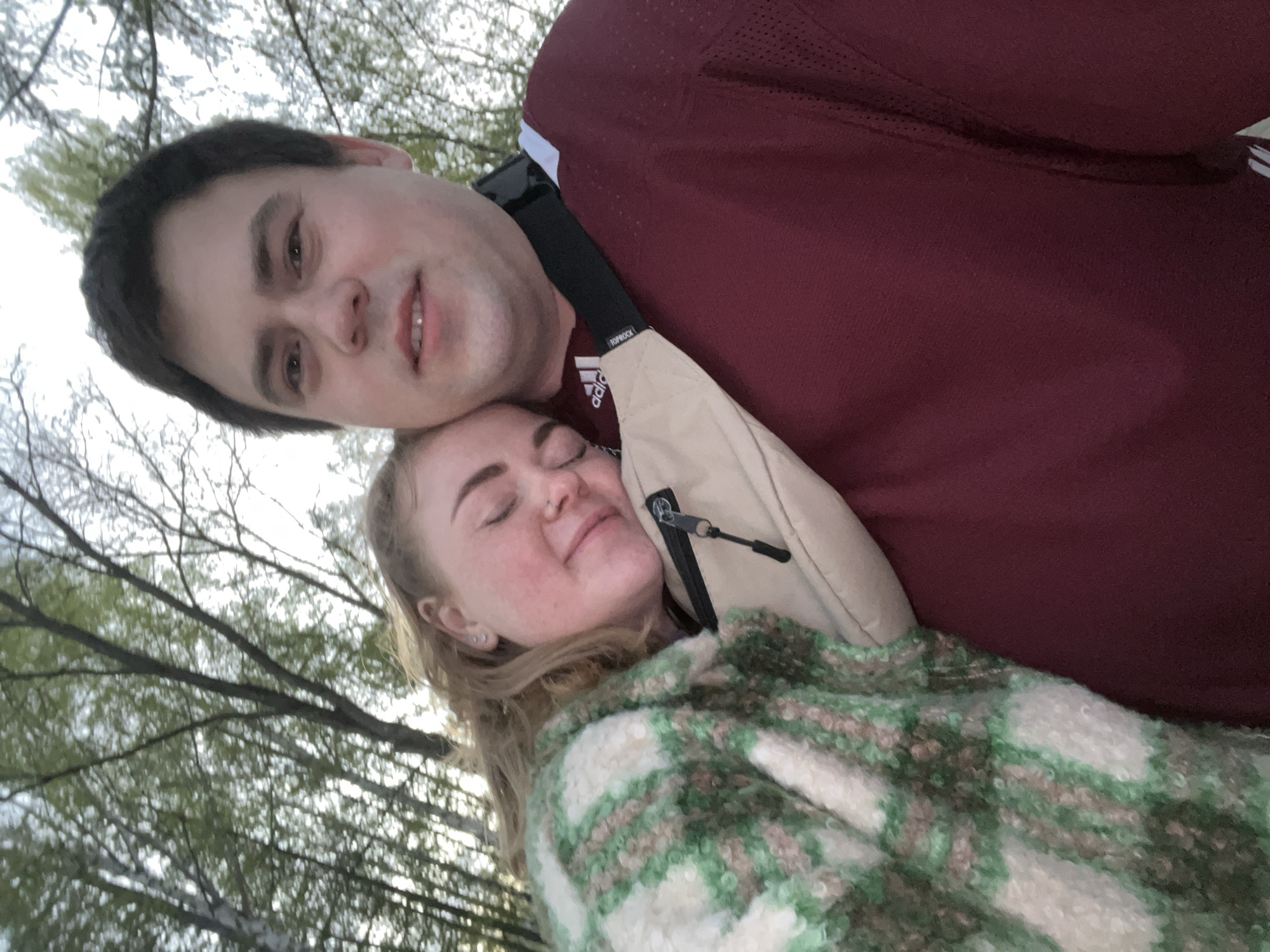 A man and a woman pose closely in an outdoor setting, likely in Chaikovsky, Russia given the prompt context. The man has short, dark hair and is wearing a maroon jersey with Adidas branding. He is looking slightly downward and to the right. The woman, who is resting her head on the man's shoulder, has fair skin, blonde hair, and closed eyes, possibly asleep. She is wearing a green and white plaid coat or blanket. A light-colored bag strap is visible across the man's torso. The background suggests a wooded area, with bare tree branches visible against the sky. The image appears to be taken at an angle.