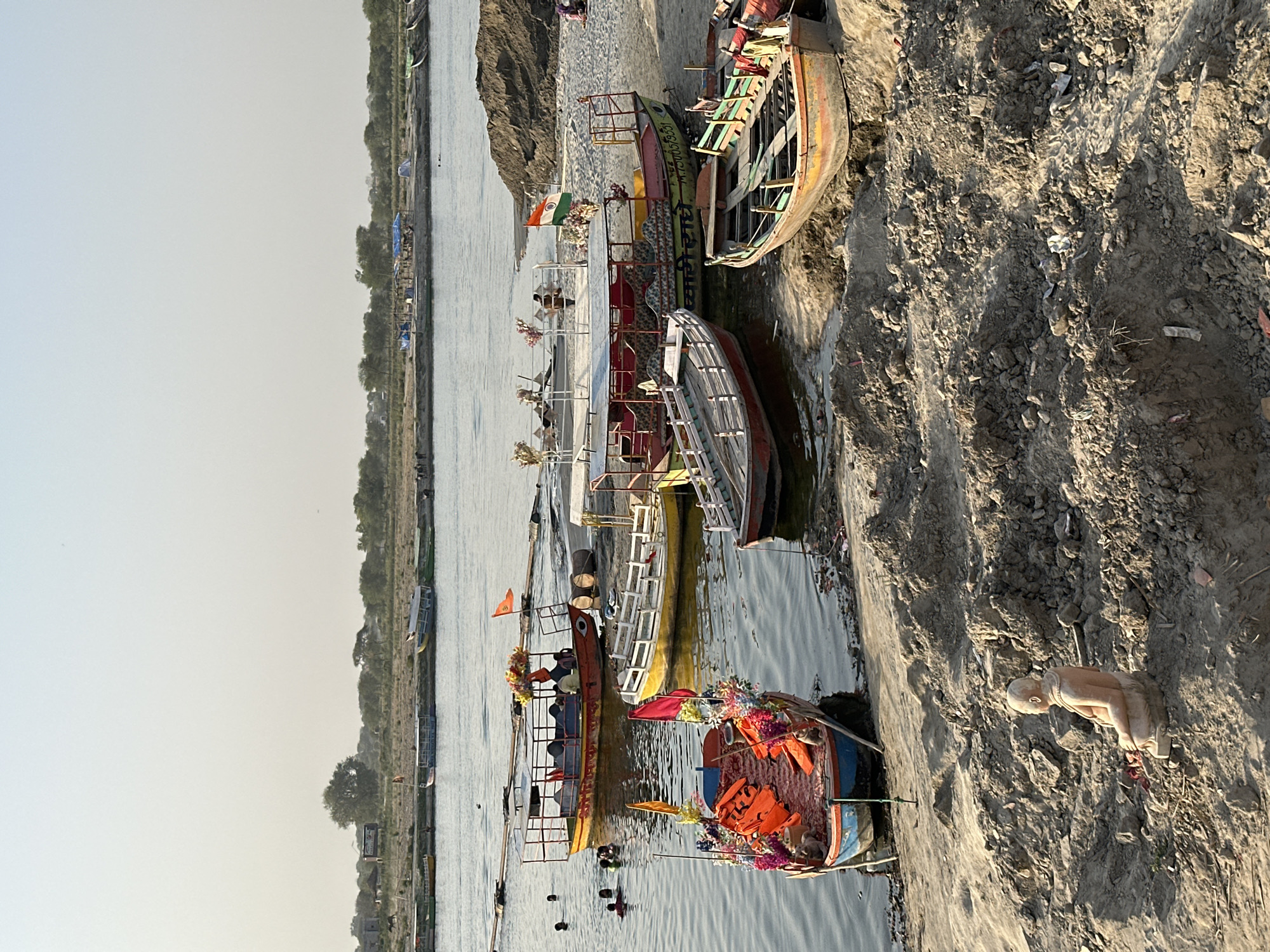 The image depicts a waterfront scene in Vrindavan, India, with a series of boats docked along a sandy bank. Several boats, ranging in size and color, are adorned with flags, decorative flowers, and life vests. Some boats have multiple levels and seating areas. People are seen swimming or standing in the water near the boats. On the sandy bank, a statue of a figure in a kneeling or bowing position is visible. The background shows trees and distant structures along the opposite bank of the river.