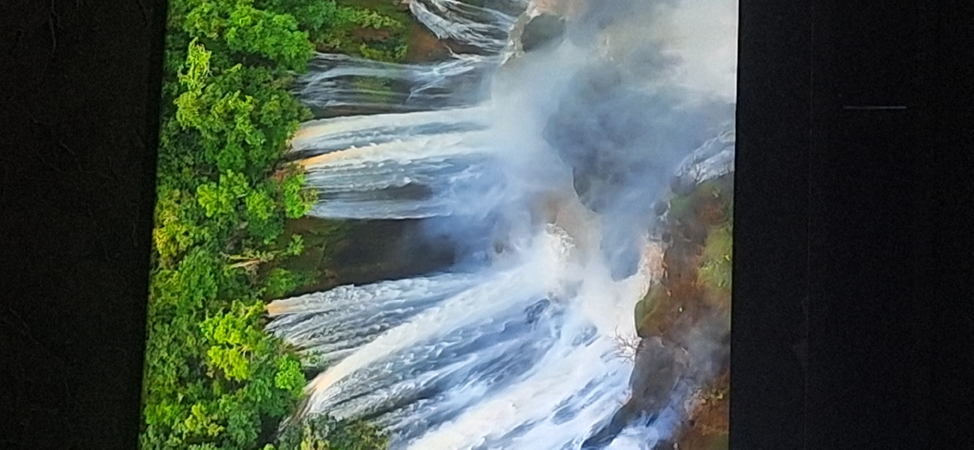 The image shows a wide shot of a lush green cliff face with multiple waterfalls cascading down. The top of the image features dense foliage, with trees and plants clinging to the rocky surface. The waterfalls are varied in width and flow, with some appearing as thin streams and others as wider curtains of water. The water crashes down into a misty pool at the bottom, creating a sense of power and dynamism. The overall scene evokes a sense of natural beauty and untouched wilderness. There is no identifiable location in the city of Bila Tserkva, Ukraine visible in this image.