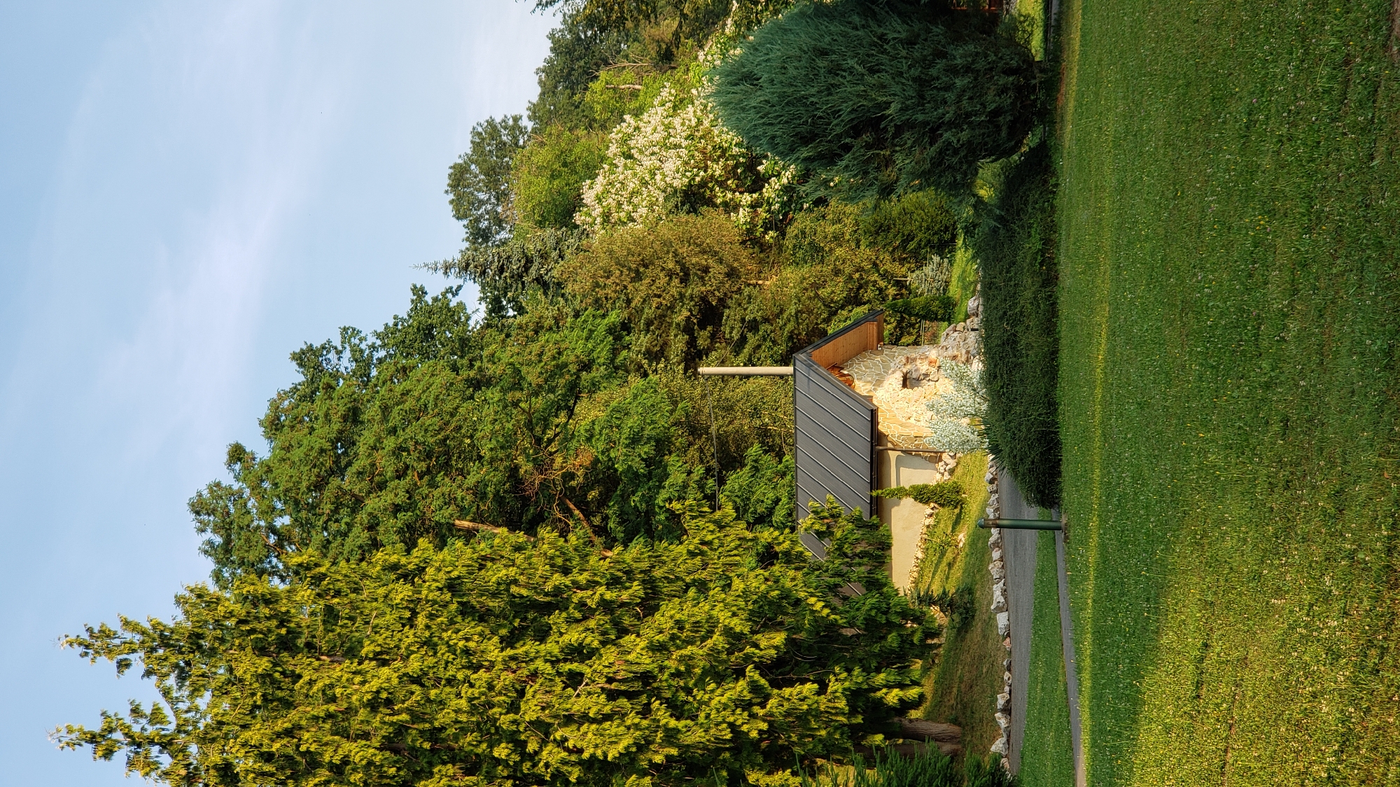 The image depicts a small, rustic building constructed with stone and a dark metal roof, situated on a grassy slope surrounded by lush green trees. A light-colored chimney extends from the roof. The building appears to be a garden structure or a small shed. In the foreground, a manicured green lawn stretches out, with a paved pathway winding through it. Several trees of varying heights and species are visible, with a prominent large evergreen on the right and a tall coniferous tree to the left of the building. The background is filled with a dense forest, and the sky is a clear blue with faint clouds. The scene suggests a tranquil outdoor setting, possibly a park or a garden. The specific location in Prešov, Slovakia, cannot be determined from the visual information provided in the image alone.