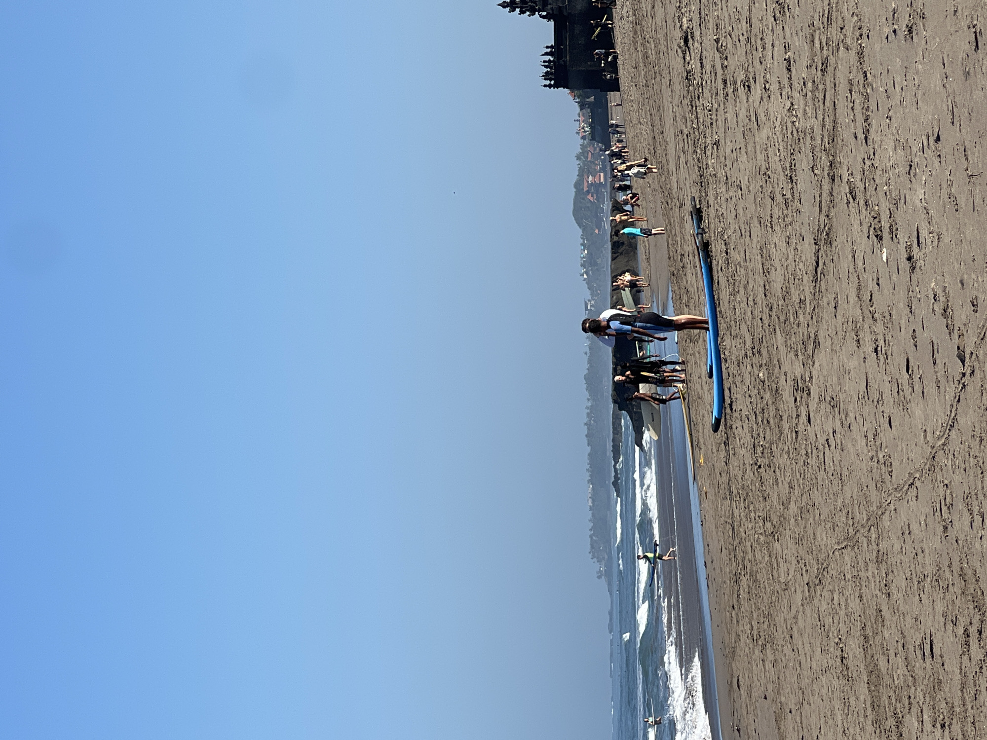 The image depicts a sunny day at a beach in North Kuta, Indonesia. The sand is light brown and textured with subtle imprints. In the foreground, a blue surfboard lies on the sand, parallel to the water's edge. Further back, a person in a black swimsuit stands next to a surfboard. The middle ground shows a group of people on the beach, some walking and others standing near the water. The ocean is a deep blue with moderate waves crashing on the shore, creating white foam. In the distance, a rocky outcrop extends into the sea, and beyond that, a coastline with scattered buildings and lush green vegetation is visible under a clear, light blue sky. To the far right of the image, a dark, ornate structure, possibly a temple, stands prominently. Several other people are dispersed along the beach and in the shallow water, some appearing to be surfers or beachgoers. The overall atmosphere is one of a lively beach scene on a bright, clear day.