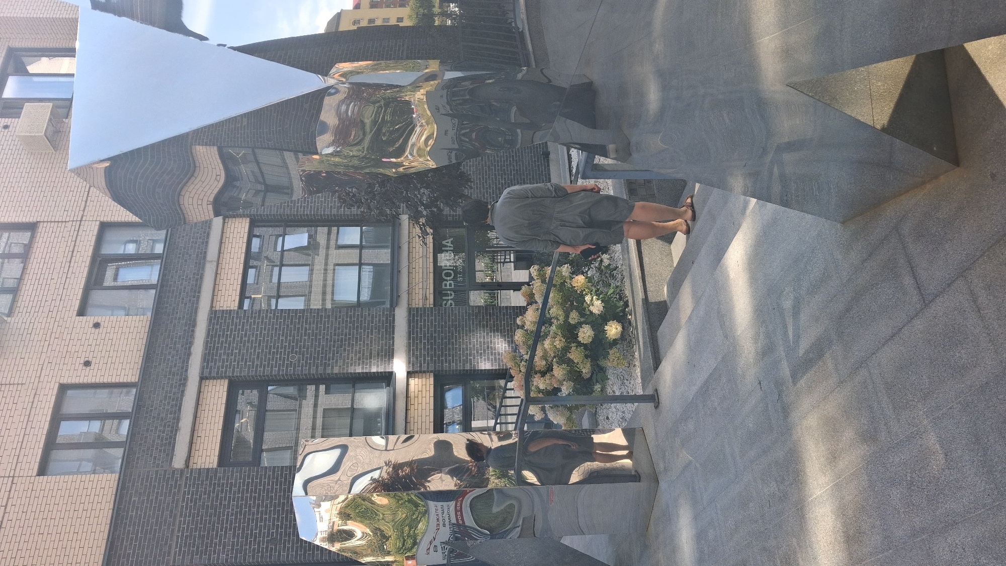 A woman with dark hair, wearing a grey jumpsuit and sandals, stands on granite steps facing away from the camera. She is looking towards a large, reflective metal sculpture. To her left, another reflective sculpture is visible, mirroring the woman and the surrounding environment. Behind the sculptures, a modern building with brick and dark paneling features windows and a sign reading "SUBORBIA ST. 202". A bush with pale flowers is located between the woman and the building. The scene appears to be an outdoor urban setting. The overall lighting suggests it is daytime with shadows cast by the sculptures and sunlight filtering through.