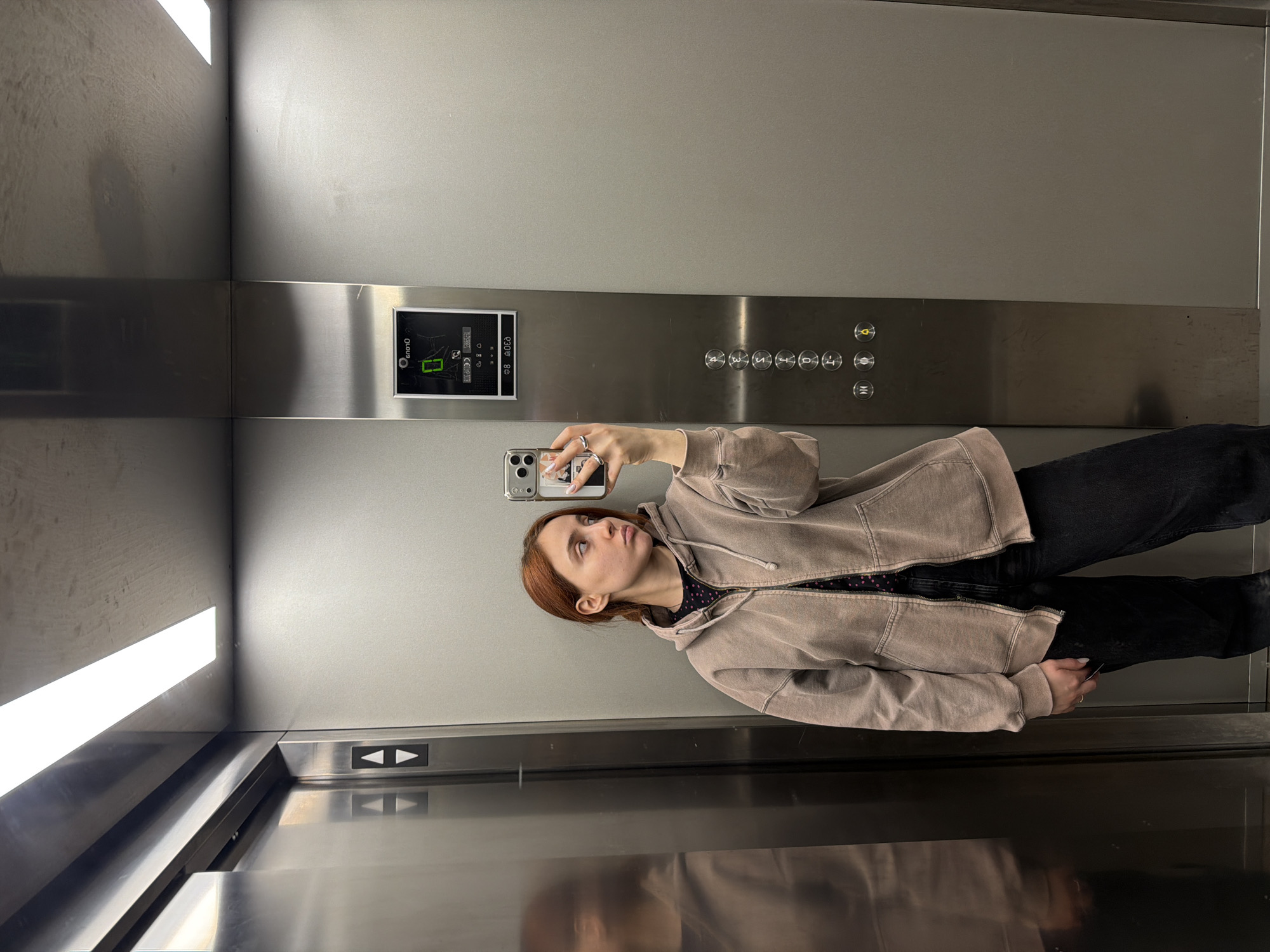 A woman with red hair is taking a selfie in an elevator. She is wearing a light brown hoodie and black pants. The elevator has silver walls and a panel of buttons labeled 0-10 and a call button. The elevator also has a lighted display above the buttons. The location is likely within Strasbourg, France, given the prompt.