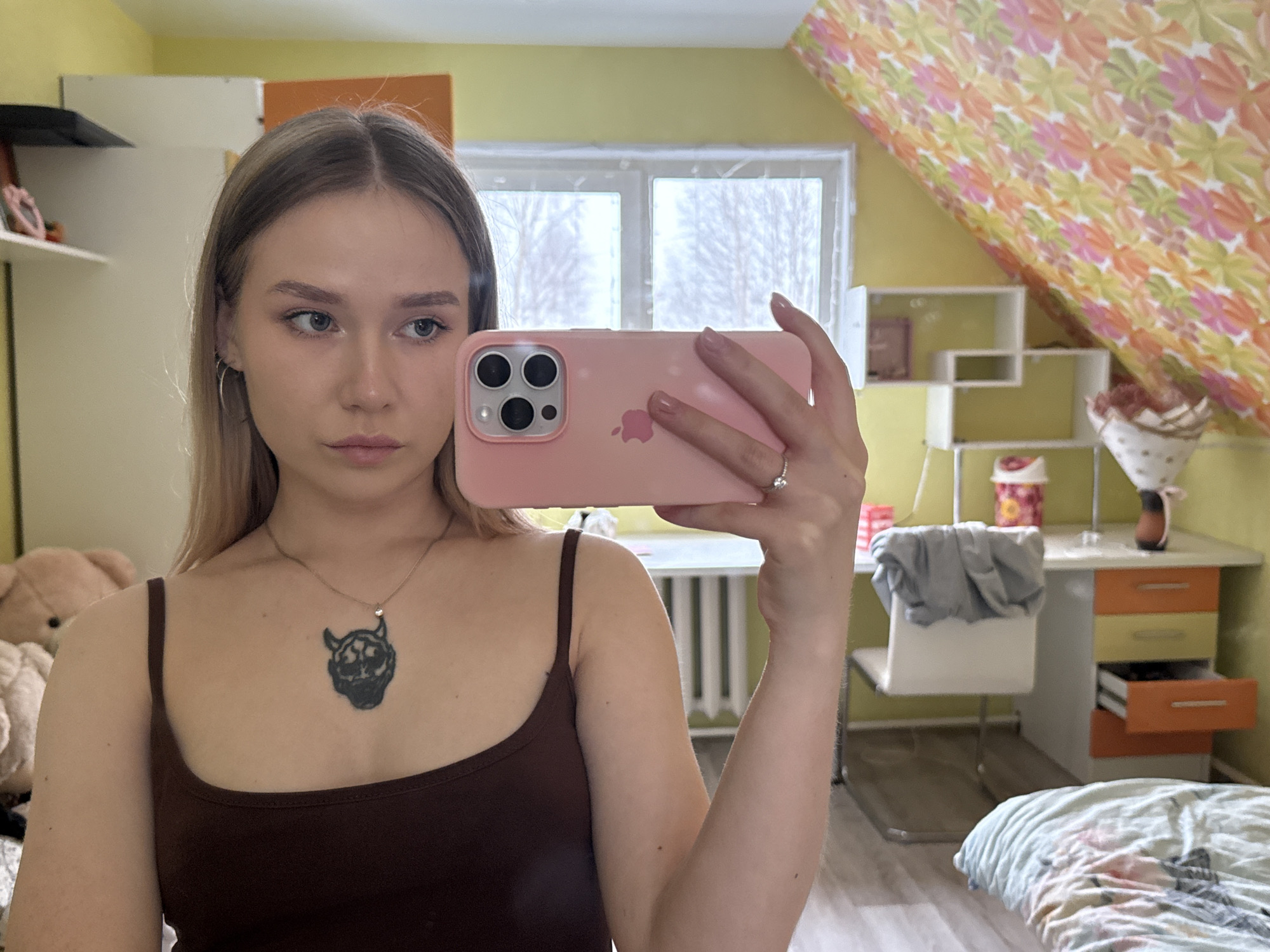 A young woman with blonde hair is taking a selfie in a room with yellow walls and a patterned ceiling, possibly in Zhigalki, Russia, though there is no immediate evidence to confirm the location. She is wearing a brown tank top and a necklace with a dark pendant. She is holding a pink iPhone, obscuring part of her face. The room contains a window with a snowy landscape visible outside, shelves, a desk with drawers, a chair, a teddy bear, and a bed with patterned bedding.