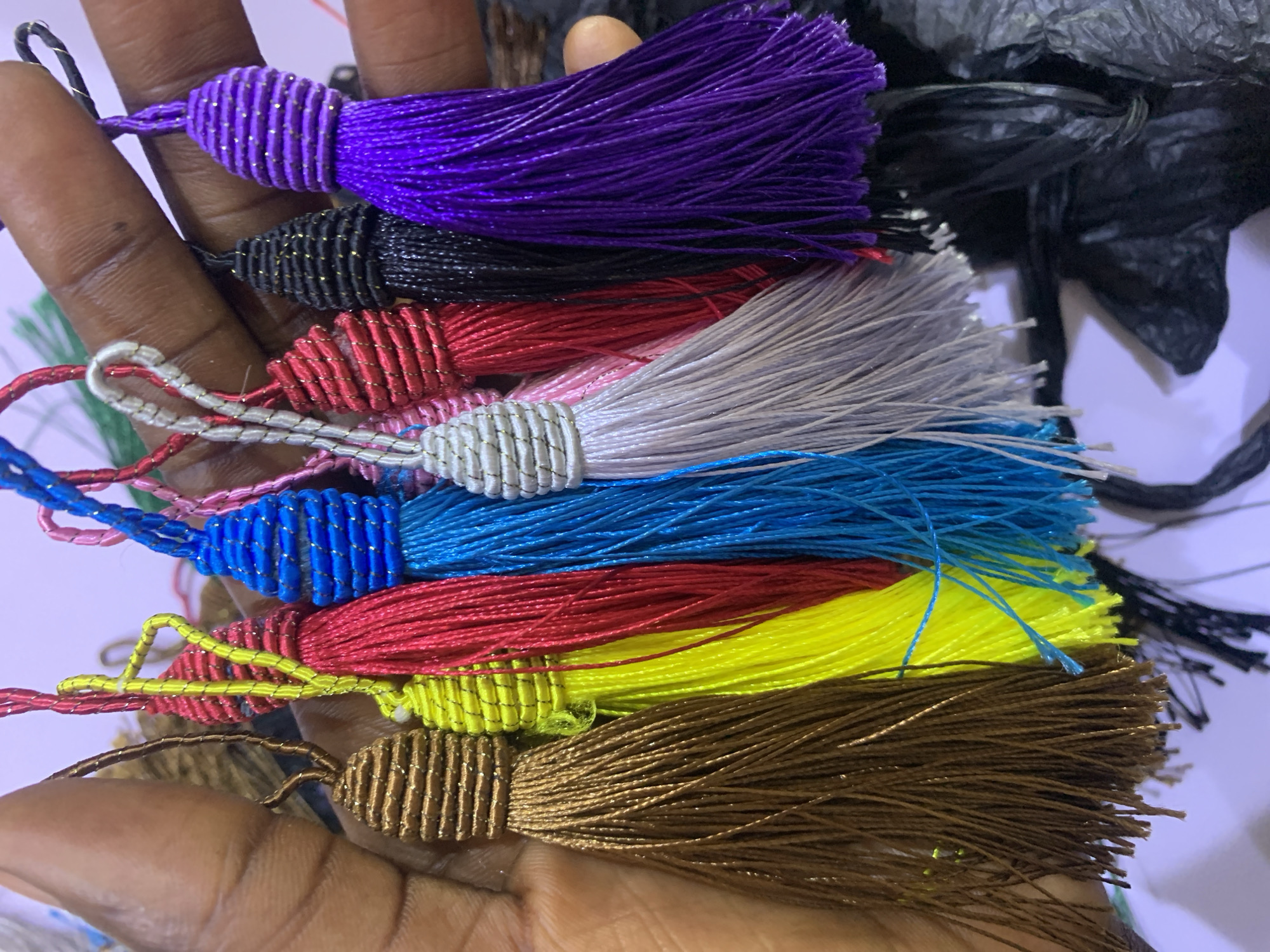 In Gura Top, Nigeria, a person's hand holds a collection of colorful tassels. The tassels, varying in hue from brown and yellow to red, blue, pink, silver, black, and purple, are made of fine, silky threads. Each tassel is topped with a decorative, ribbed cap, also crafted from thin, metallic-looking threads. The background is out of focus, suggesting a casual setting within the city.