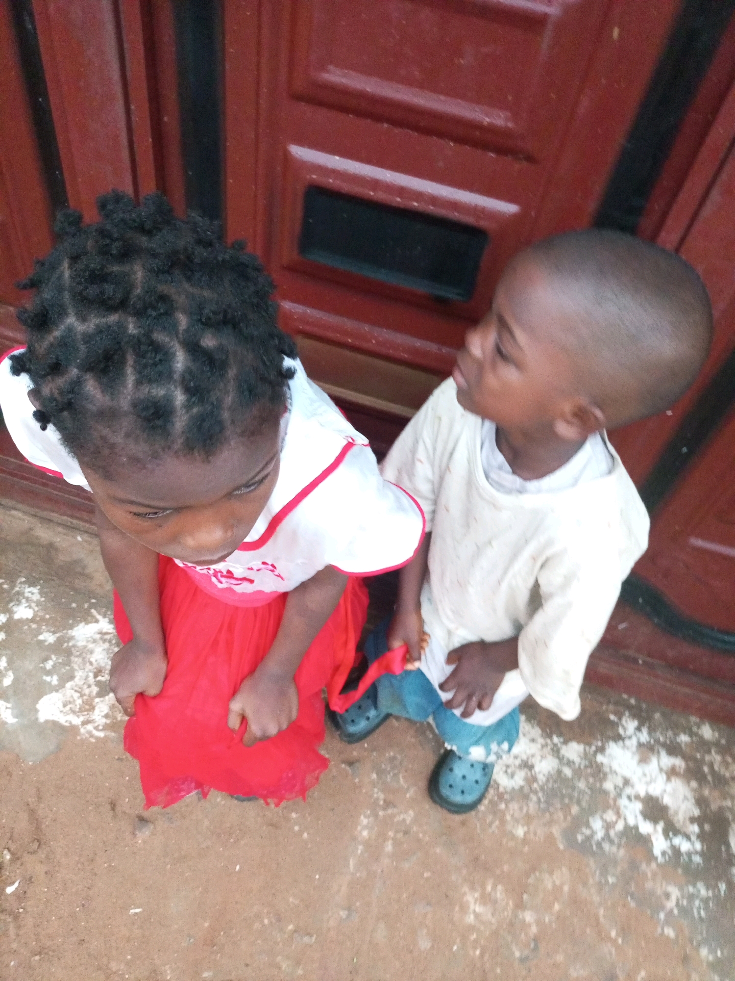 This image depicts two young children standing on a rough, dirt and concrete ground in front of a red door. The child on the left, a girl, has dark, braided hair and is wearing a white top with red trim and a vibrant red skirt. Her hands are clasped in front of her. The child on the right, a boy, has a shaved head and is wearing a light-colored, possibly cream, t-shirt and blue pants. He has on black clogs. The door behind them is painted red and has vertical paneling. The ground shows patches of white debris. The scene appears to be an outdoor setting, possibly a residential entrance, in Tempure, Nigeria.